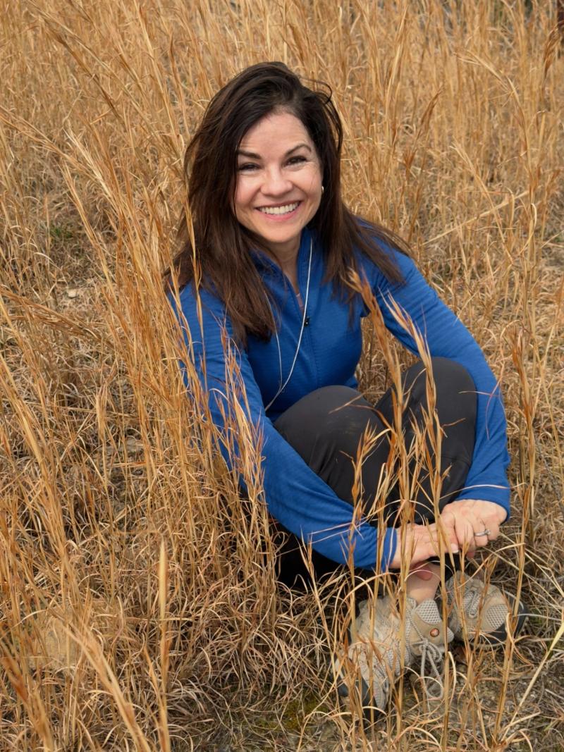 woman with dark hair smiling while seated in tall golden grass wearing a blue jacket outdoors