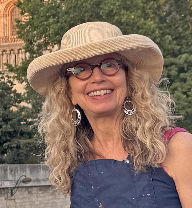 woman with curly blonde hair wearing glasses and a wide-brim hat smiling outdoors