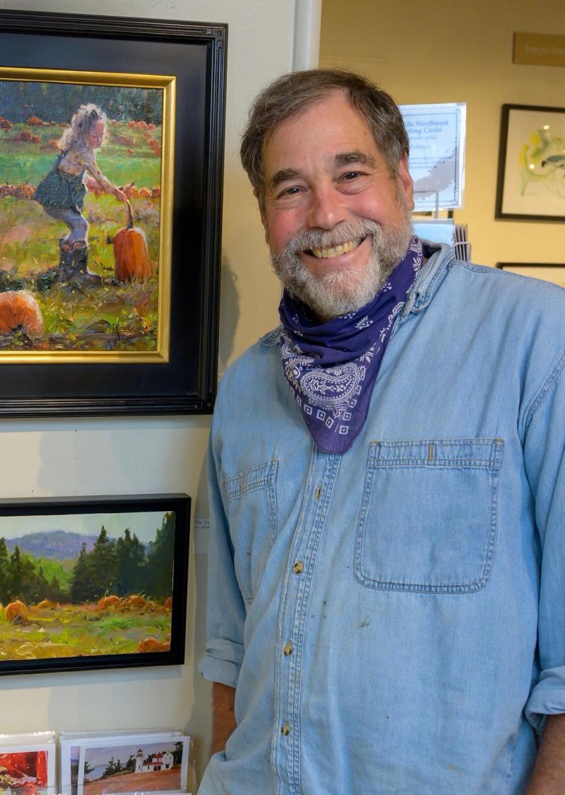 smiling gray-bearded man in a denim shirt and bandana in an art gallery