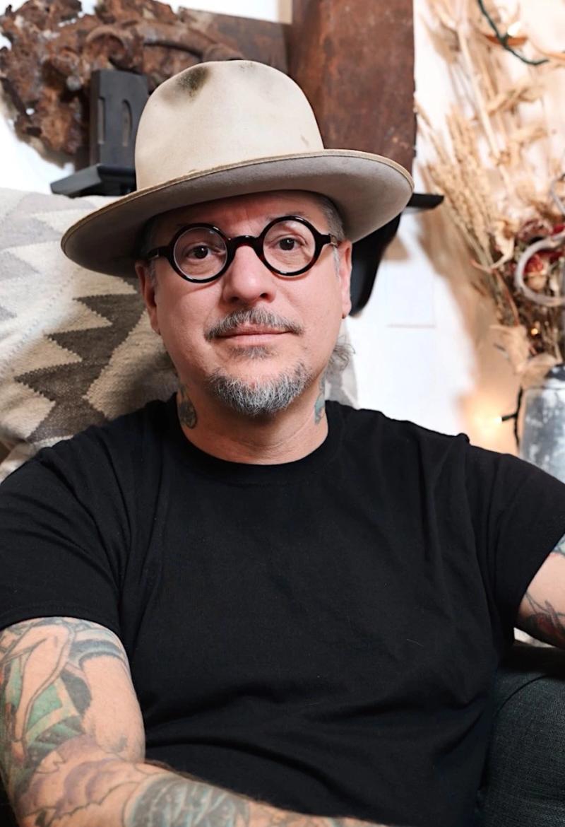 portrait of a man wearing round glasses, a tan brimmed hat, and a black t-shirt with tattooed arms