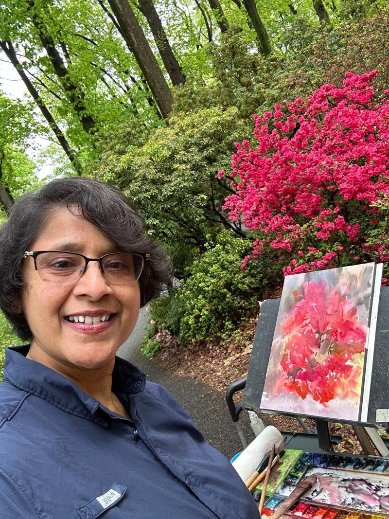 woman with glasses smiling outdoors beside an easel with a watercolor painting of bright pink azaleas