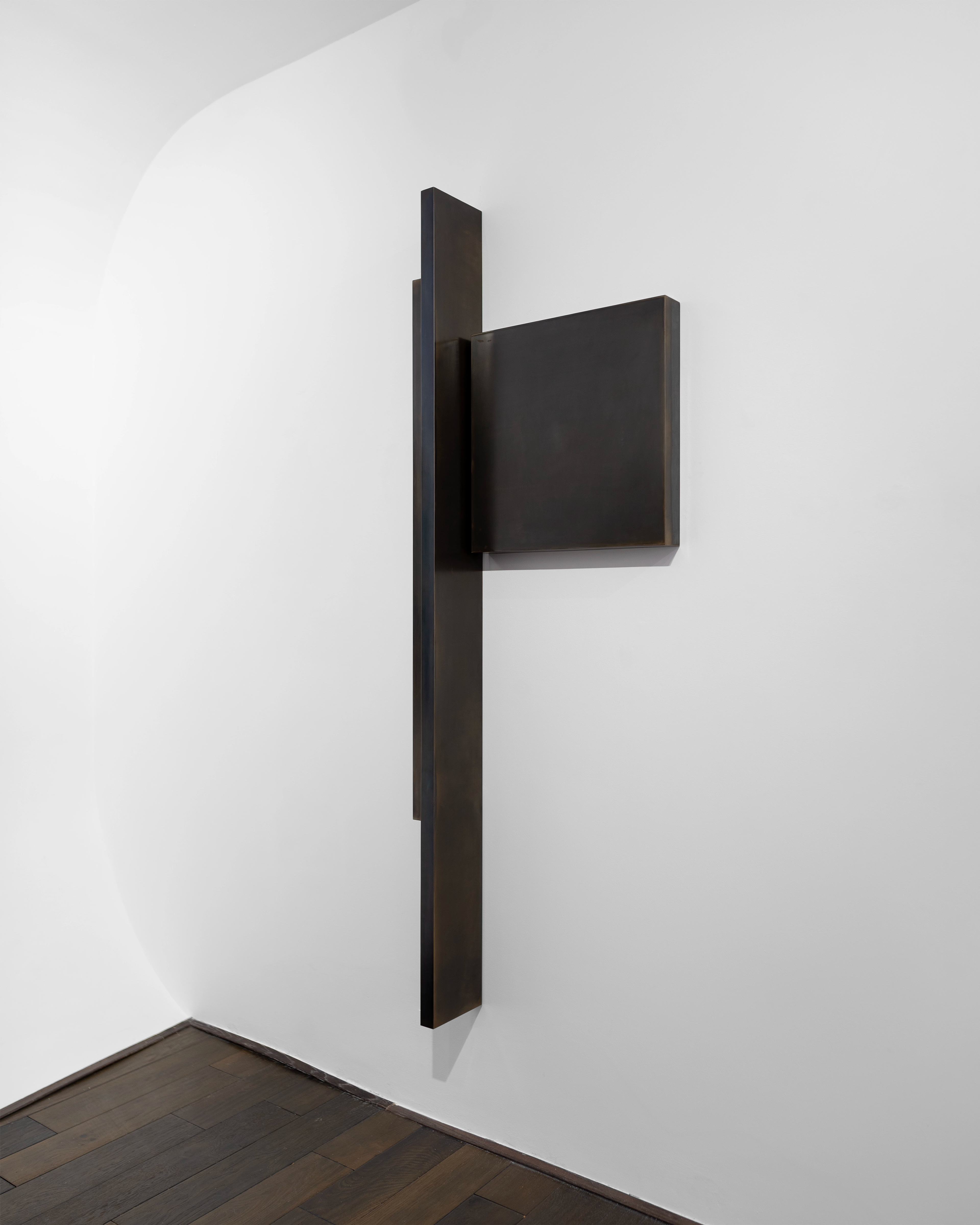 Vertical dark steel structure mounted on white wall, with a rectangular protruding element, casting sharp shadow
