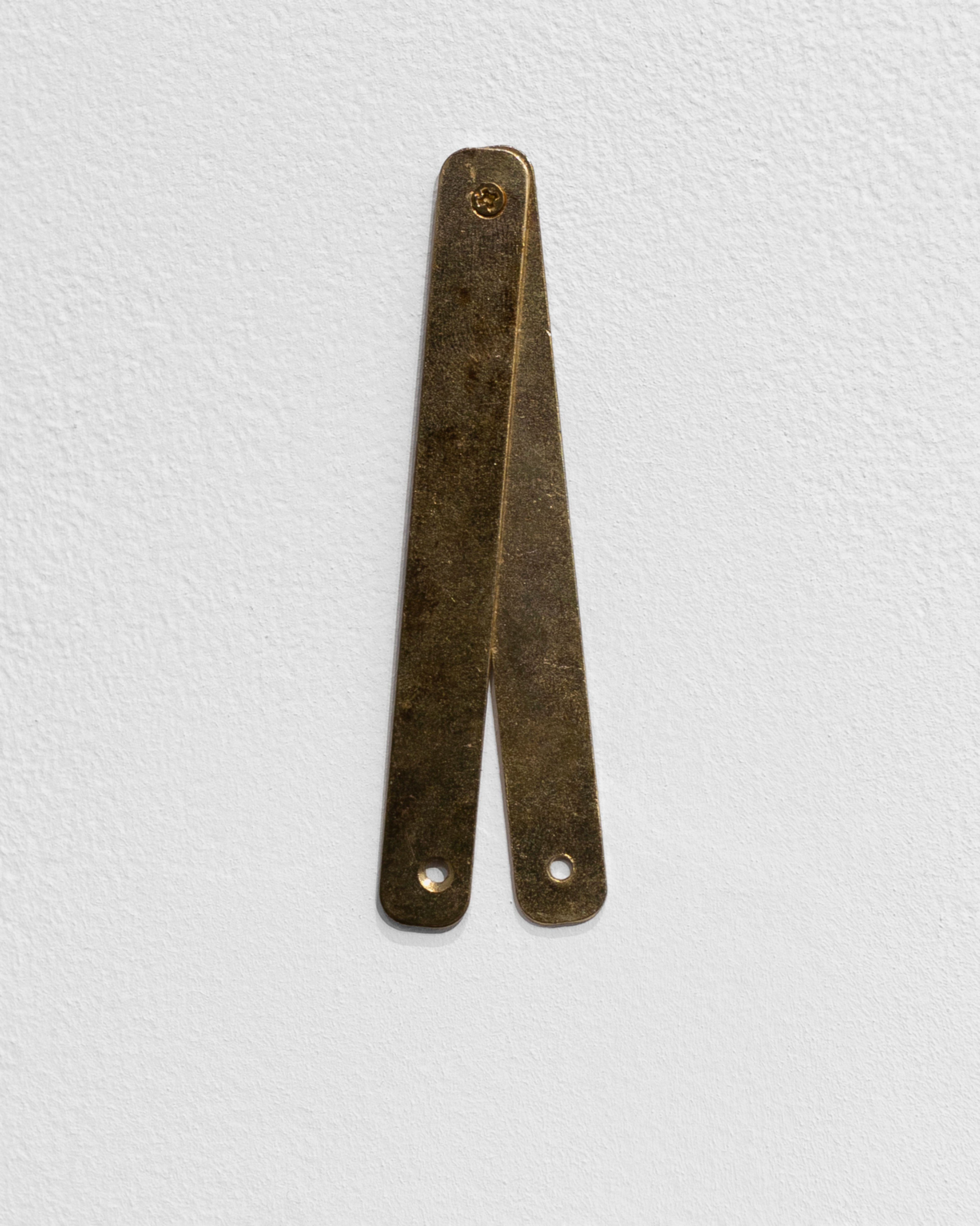 Patinated brass scissor-like form with split lower legs and circular apertures, mounted vertically on white wall