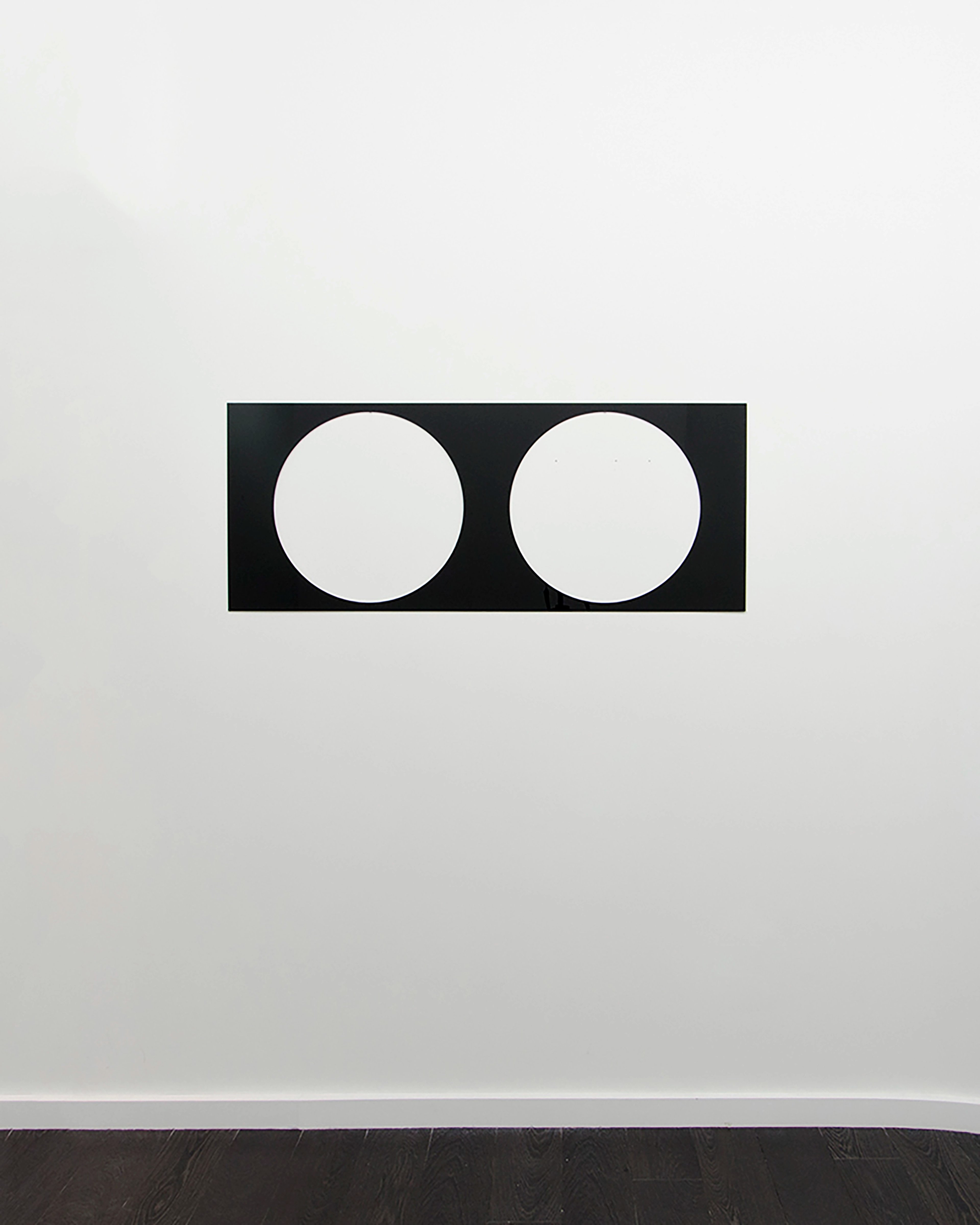 Black rectangular panel with two white circles cut through, mounted on white wall above dark wooden floor