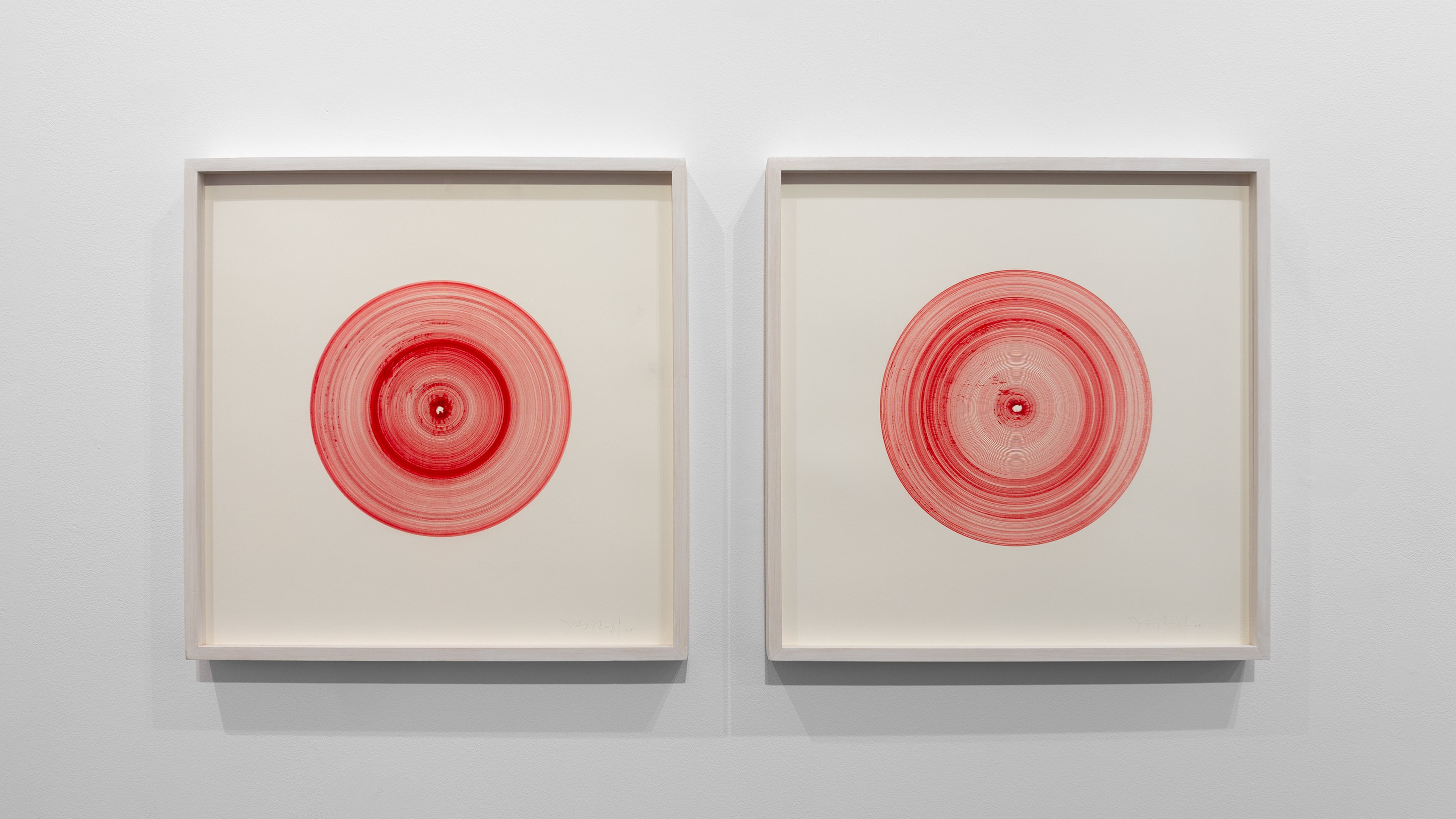 Two framed drawings with concentric red circles on cream paper, each centered within white frames, hung side by side on a gallery wall