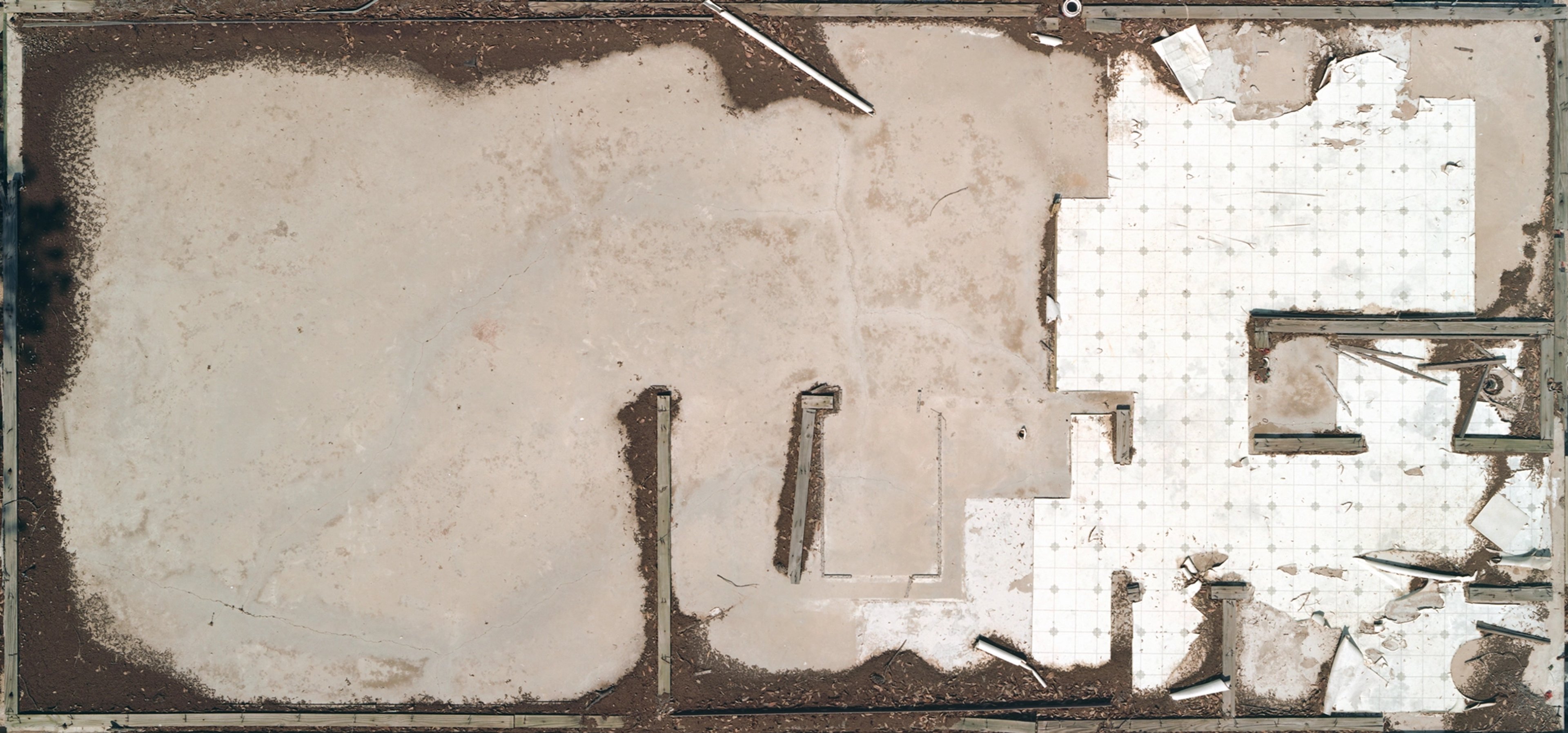 Overhead photograph of a derelict room with peeling walls, two vertical pipes, scattered white paper debris, and shadowed corners in muted beige and brown to…