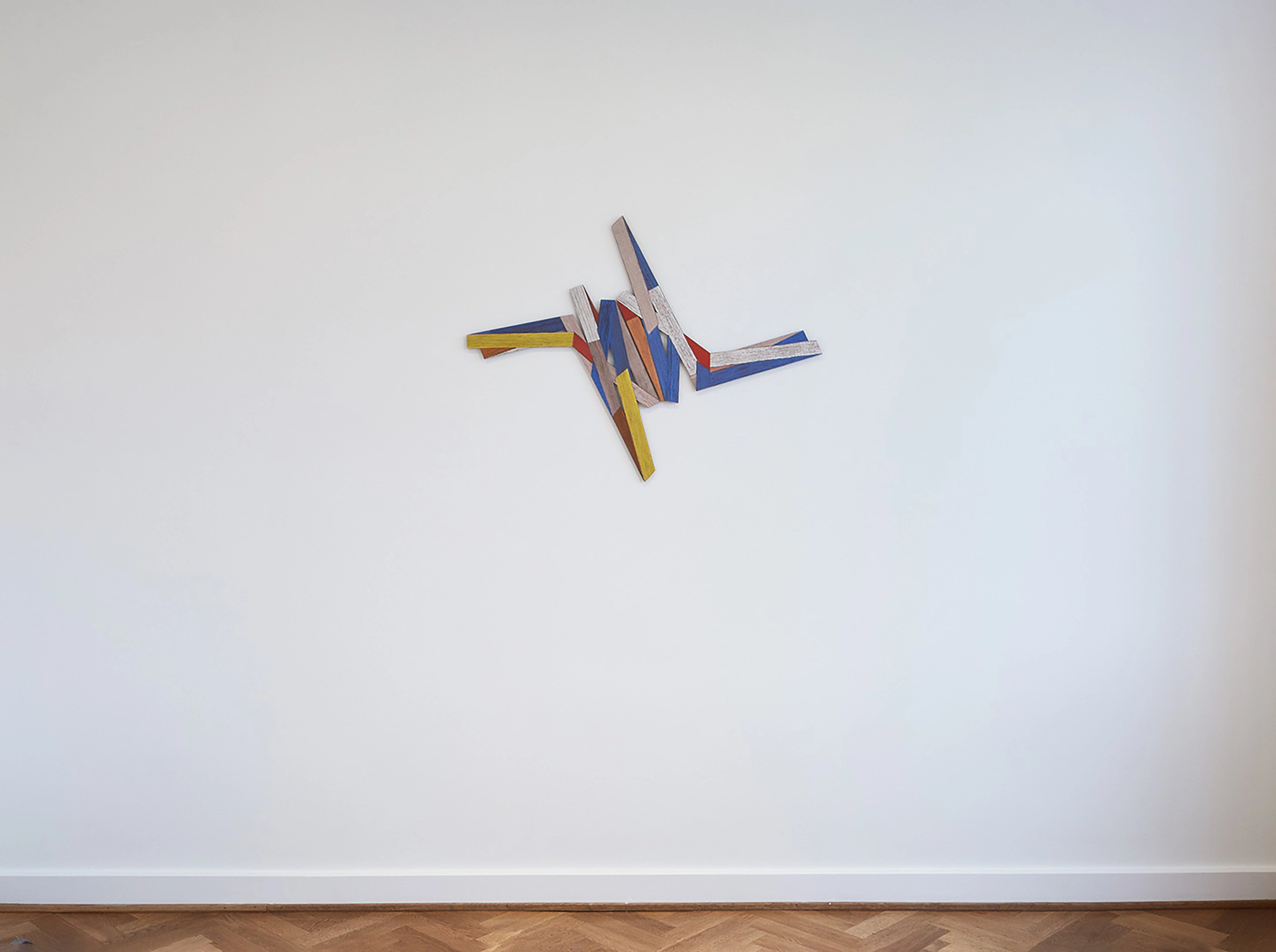 Painted wooden rods in yellow, blue, grey, and red arranged in a dynamic angular composition against a white wall
