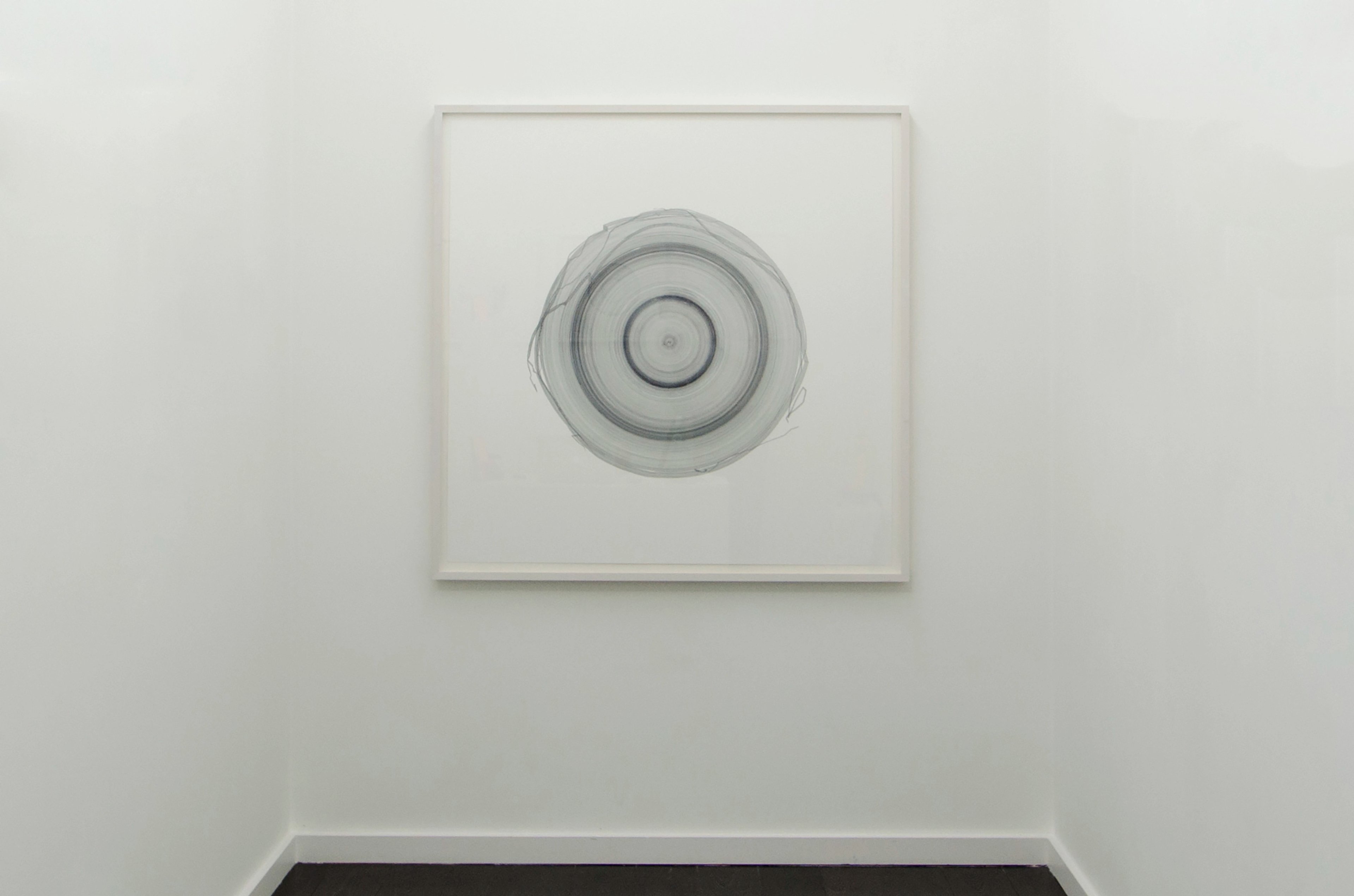 Concentric circular forms drawn in graphite grey pigment on cream paper, mounted in a light frame on white gallery wall