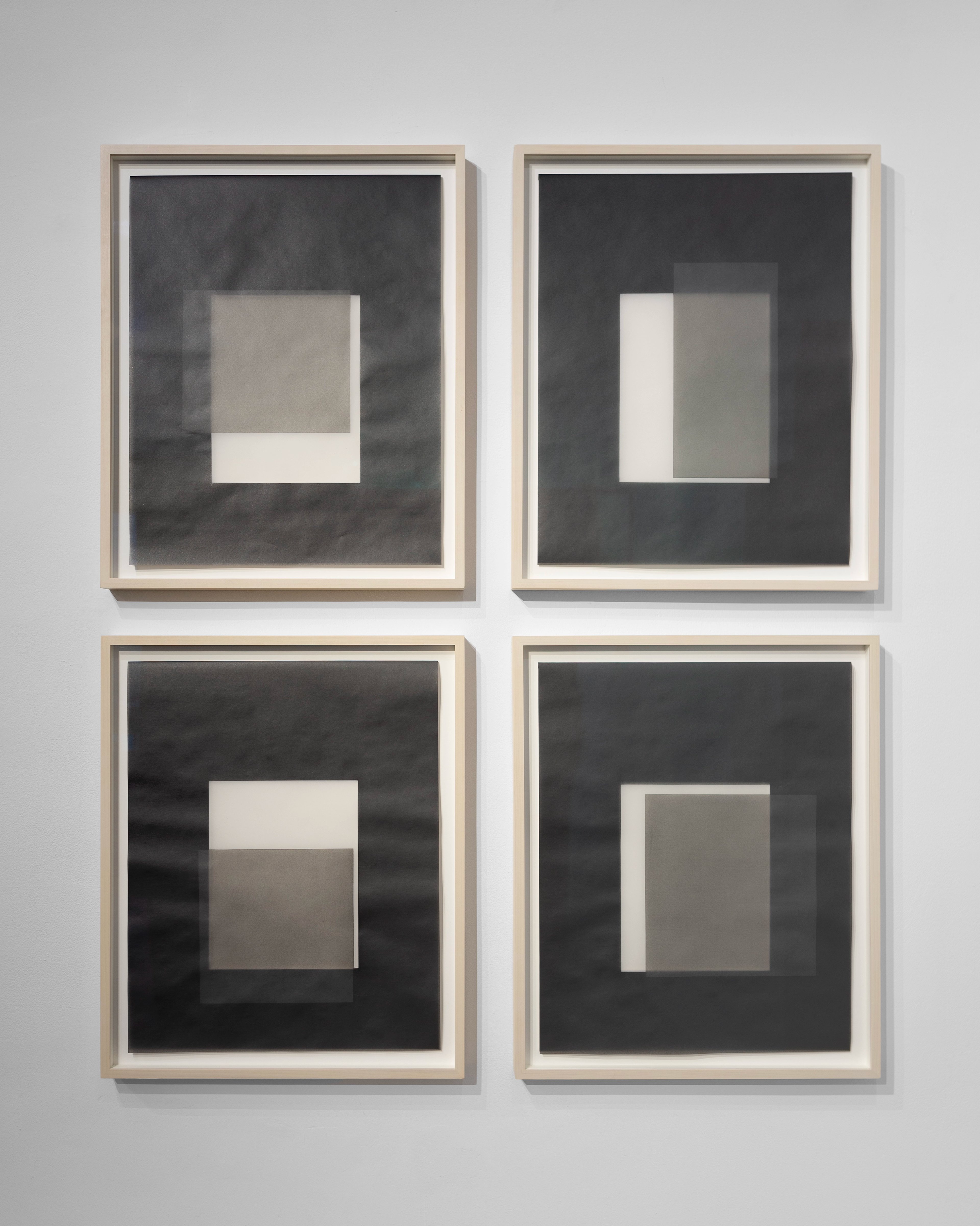 Four framed works on paper in light wood frames arranged in a grid, each featuring layered geometric rectangles in white, grey, and charcoal tones on dark ba…