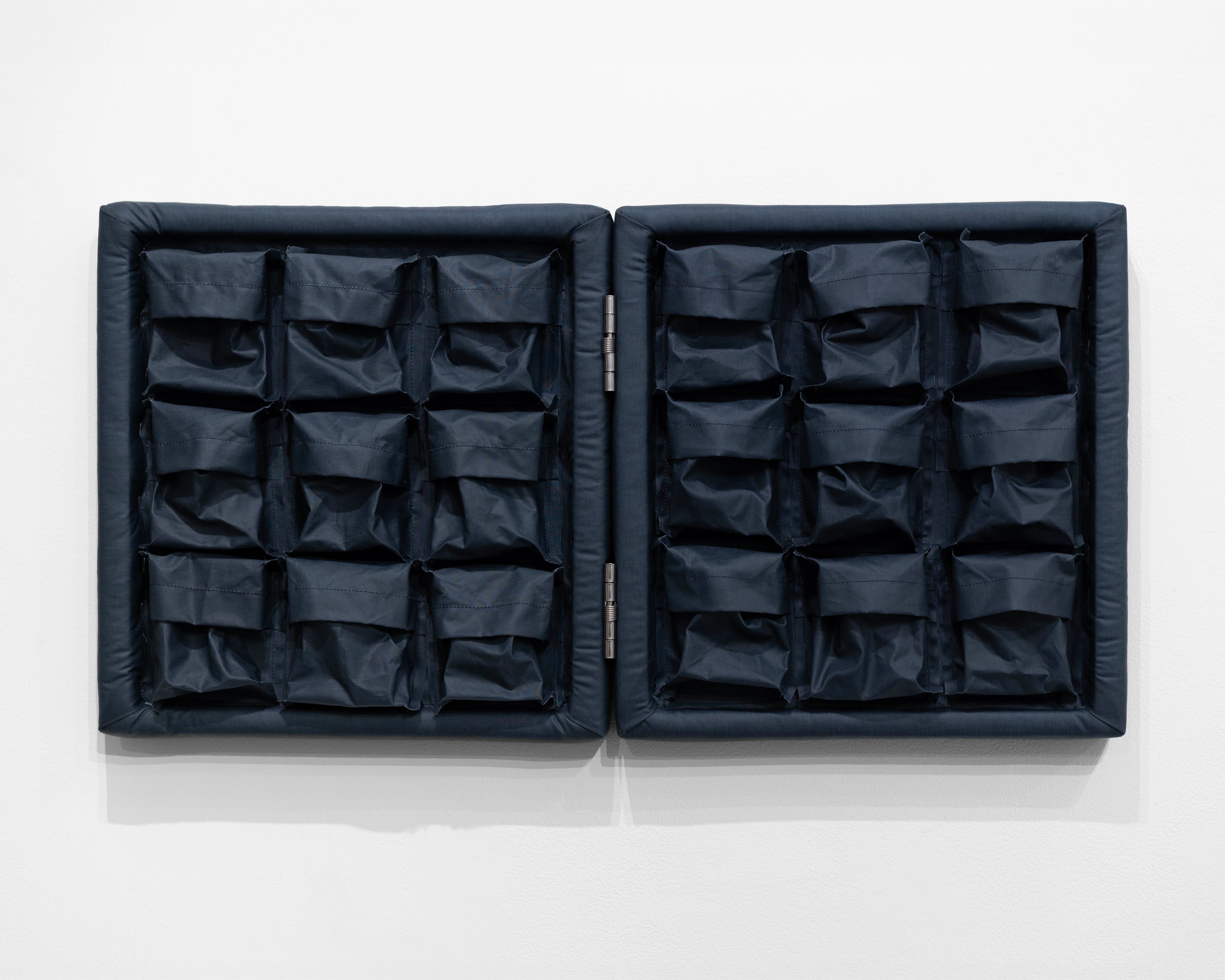 Two adjoining dark navy oilskin panels, each divided into nine quilted pockets arranged in a 3x3 grid, mounted horizontally on white wall