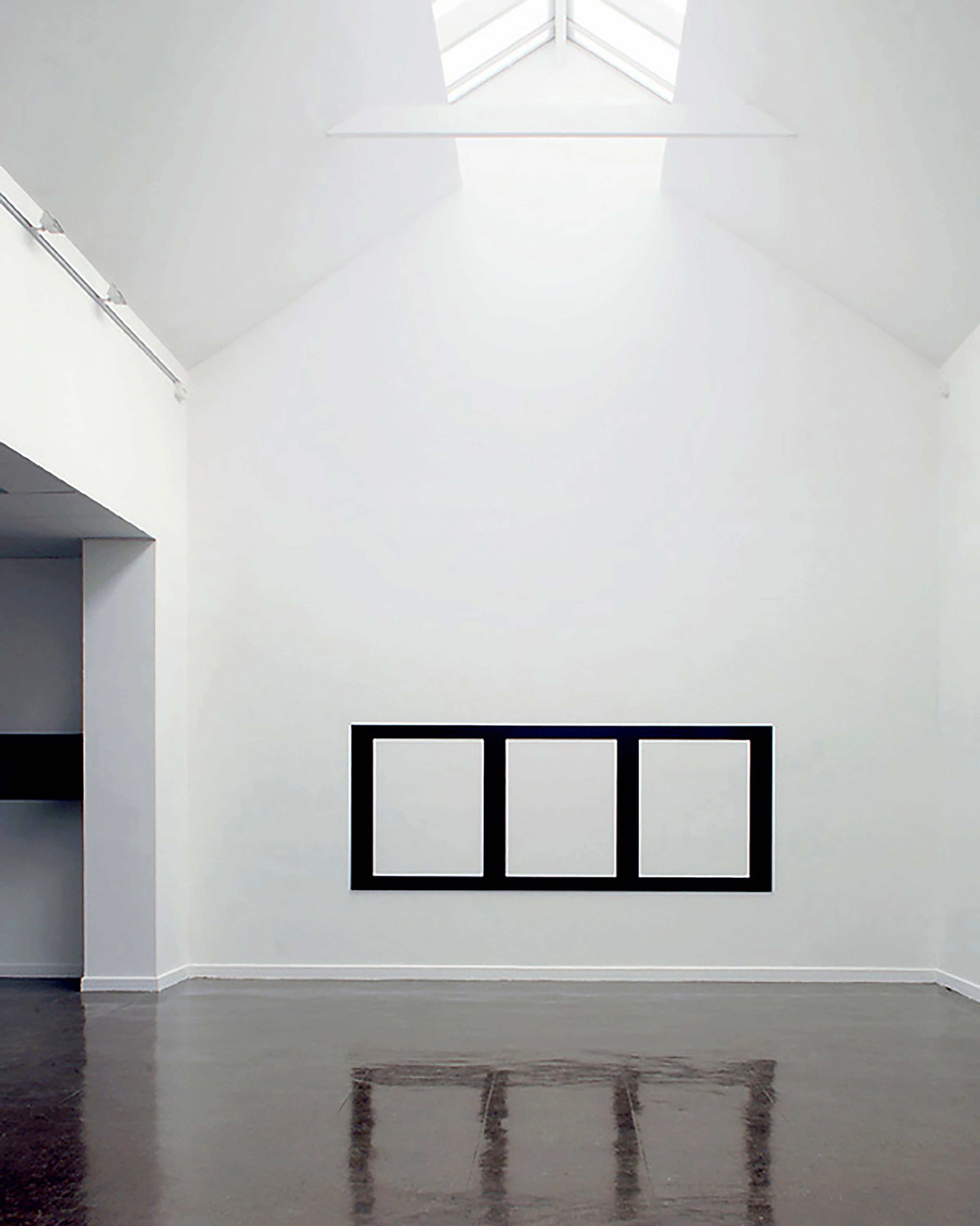Three black-framed rectangular openings mounted horizontally on a white gallery wall, with reflective surfaces below on dark flooring beneath a skylit ceiling