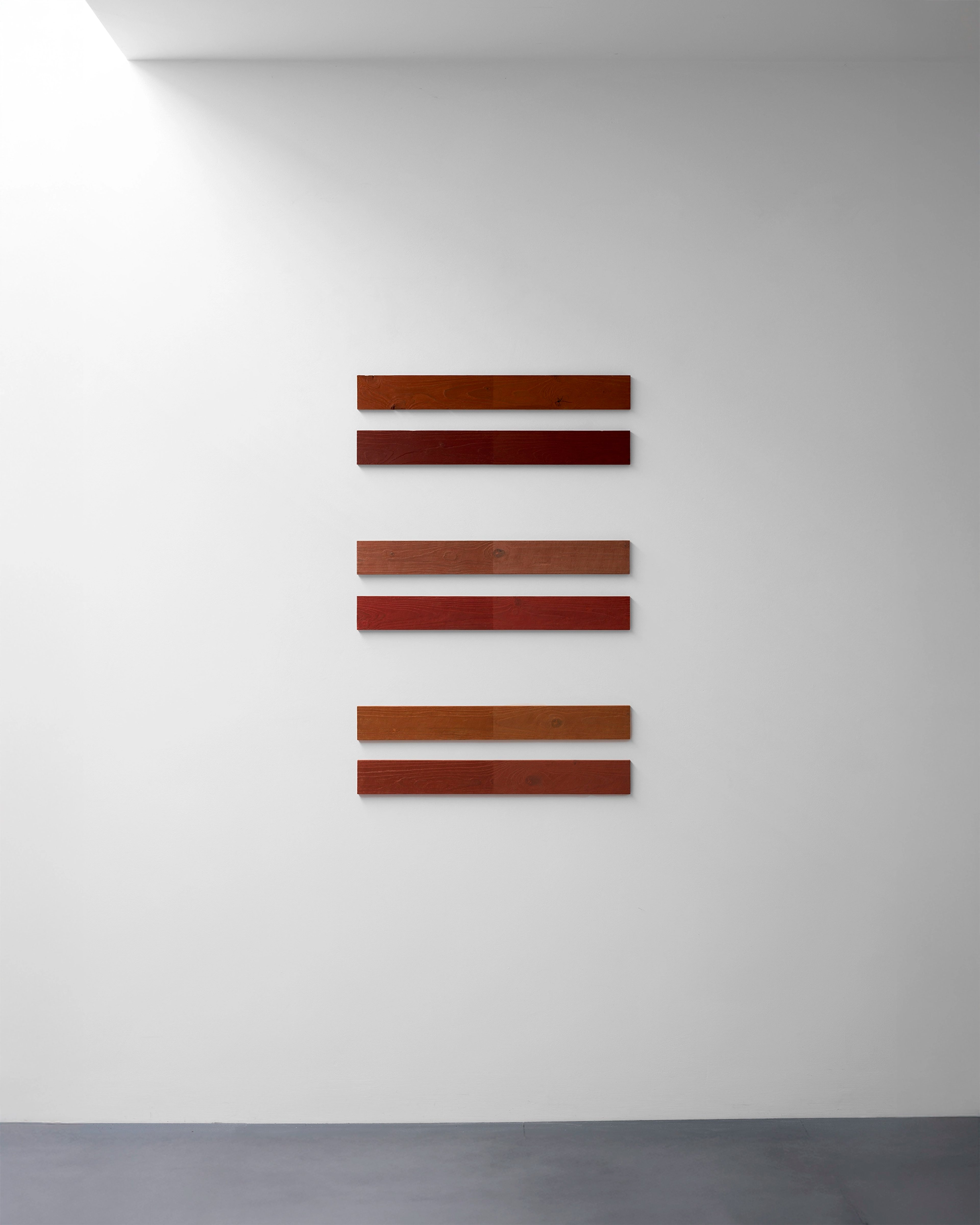 Six horizontal wooden bars in alternating brown and deep red tones arranged in three pairs on a white gallery wall, created from reclaimed wood and wax pencil