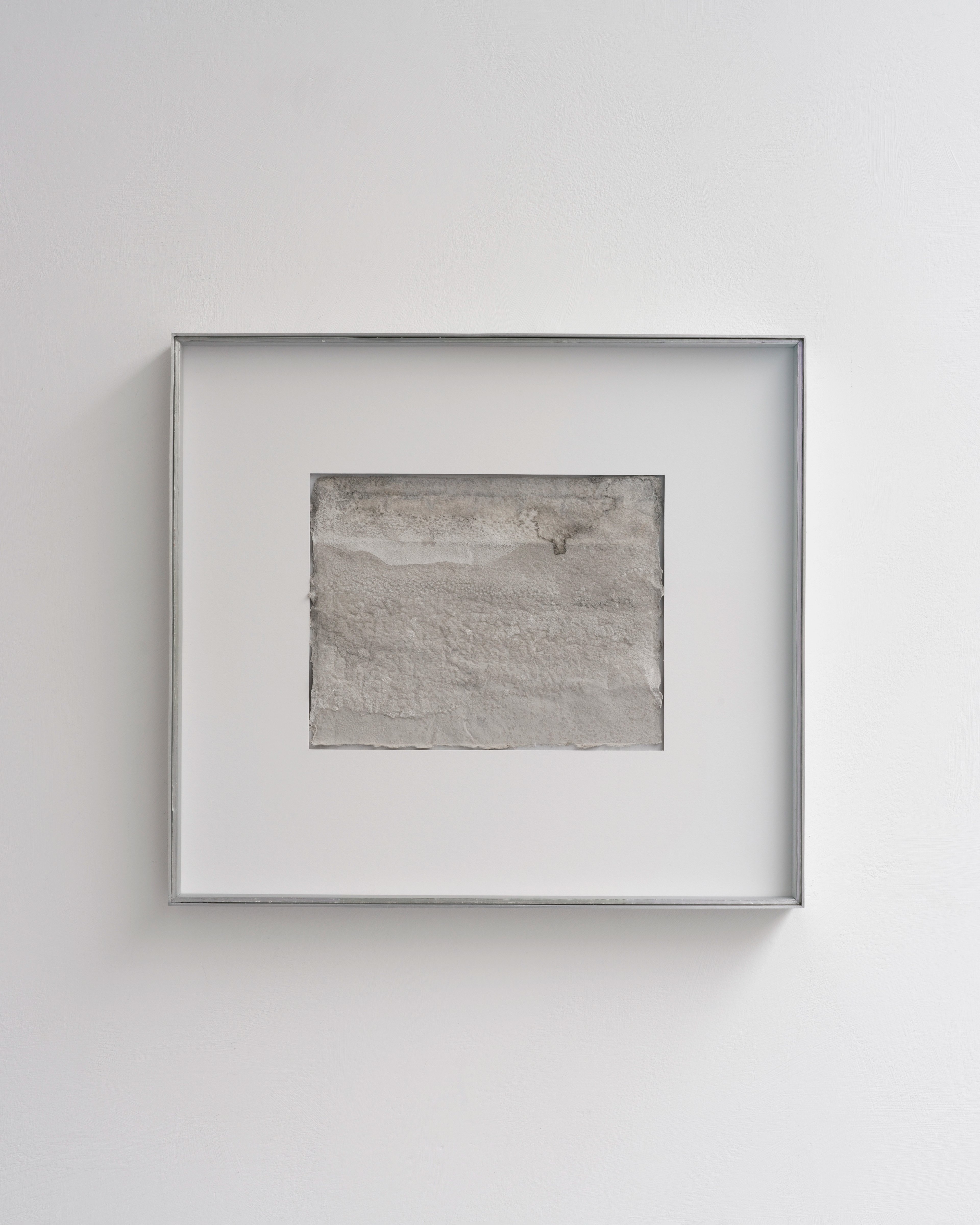 Framed handmade Japanese paper with textured grey-brown surface created using ink and seawater, mounted centrally on white mat within silver frame
