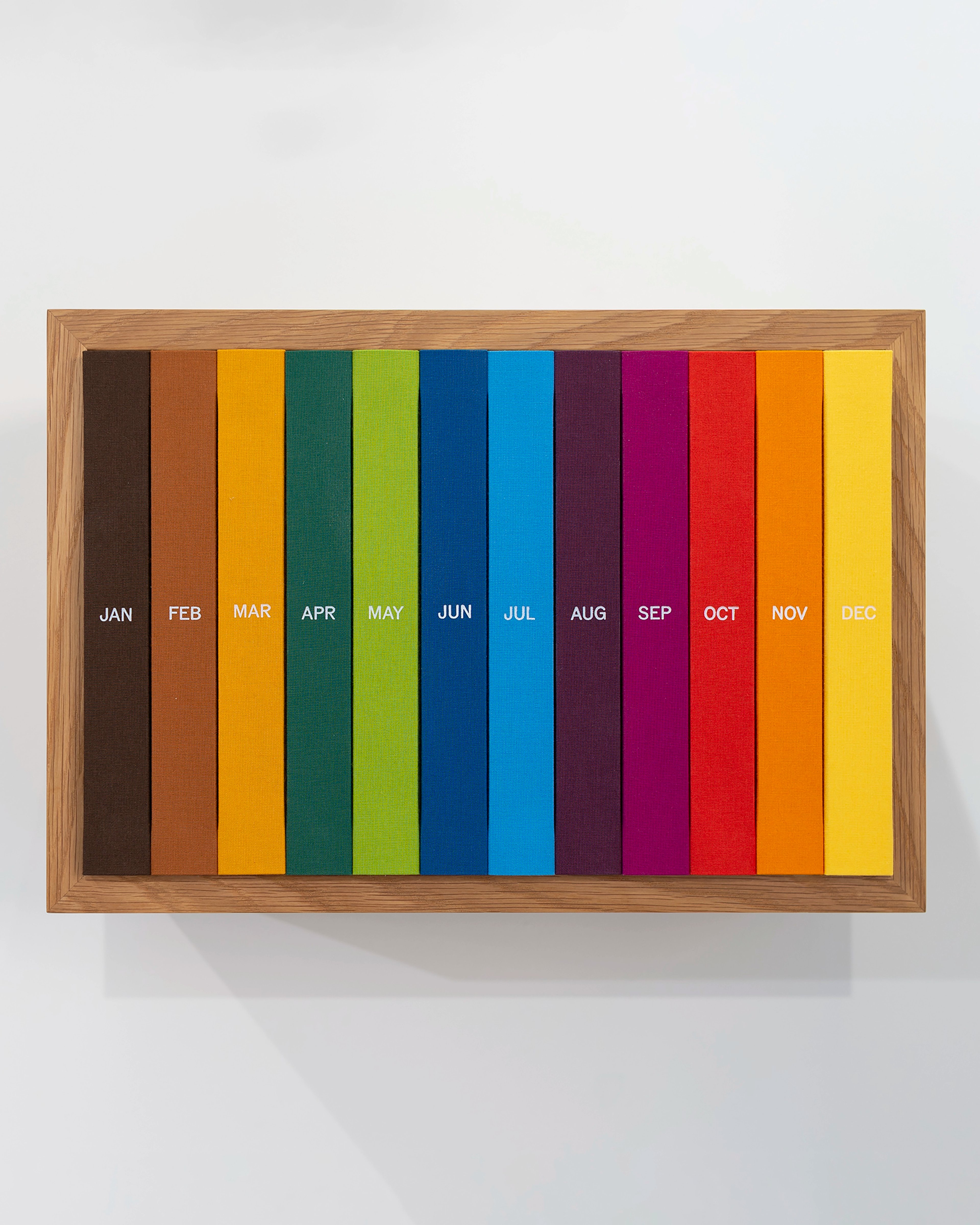 Twelve vertically stacked colored paper spines labeled with month abbreviations, arranged chronologically from brown January through yellow December, mounted…