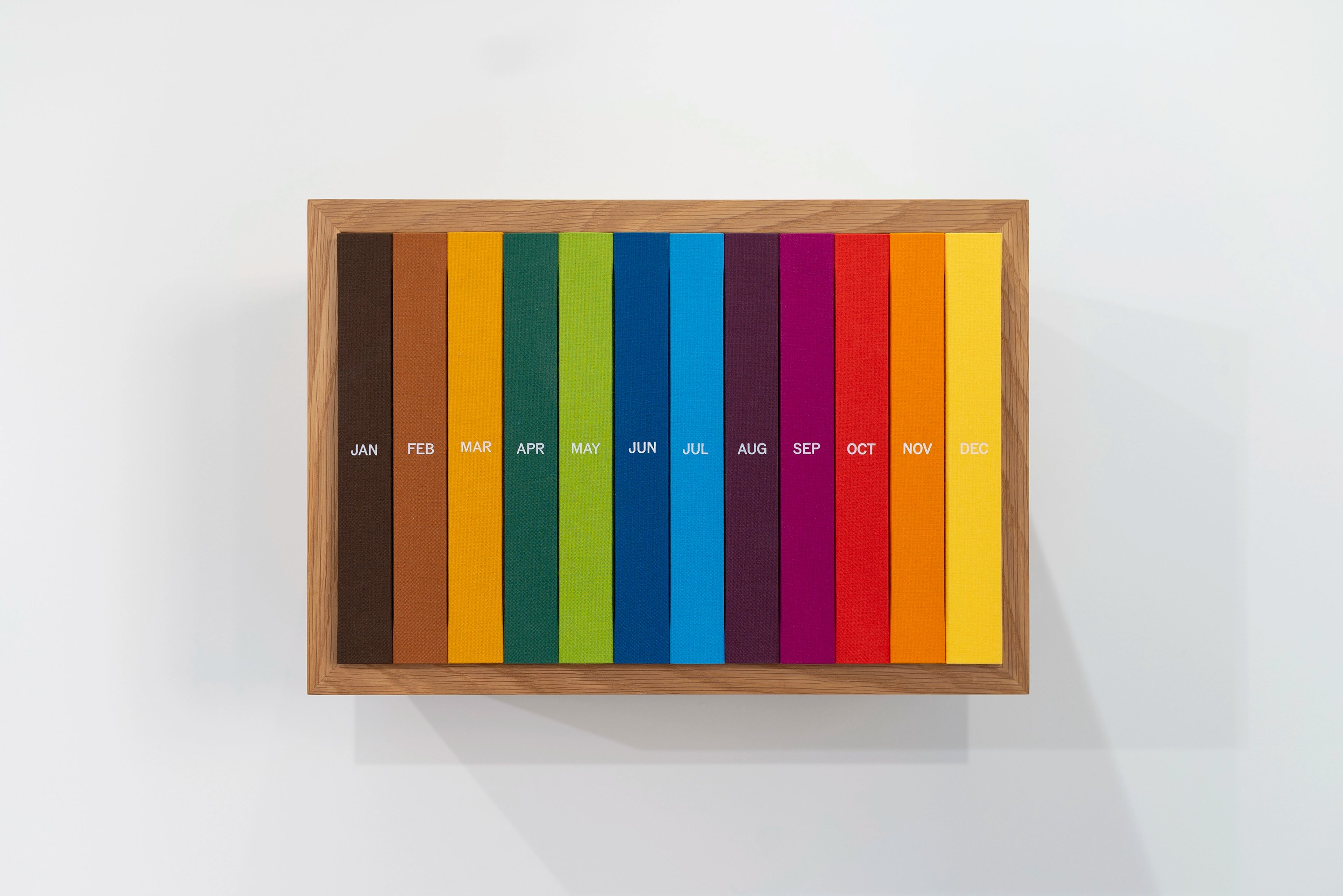 Twelve vertical colored paper strips mounted in a wooden frame, each labeled with a month abbreviation, arranged in spectral order from brown through red, or…