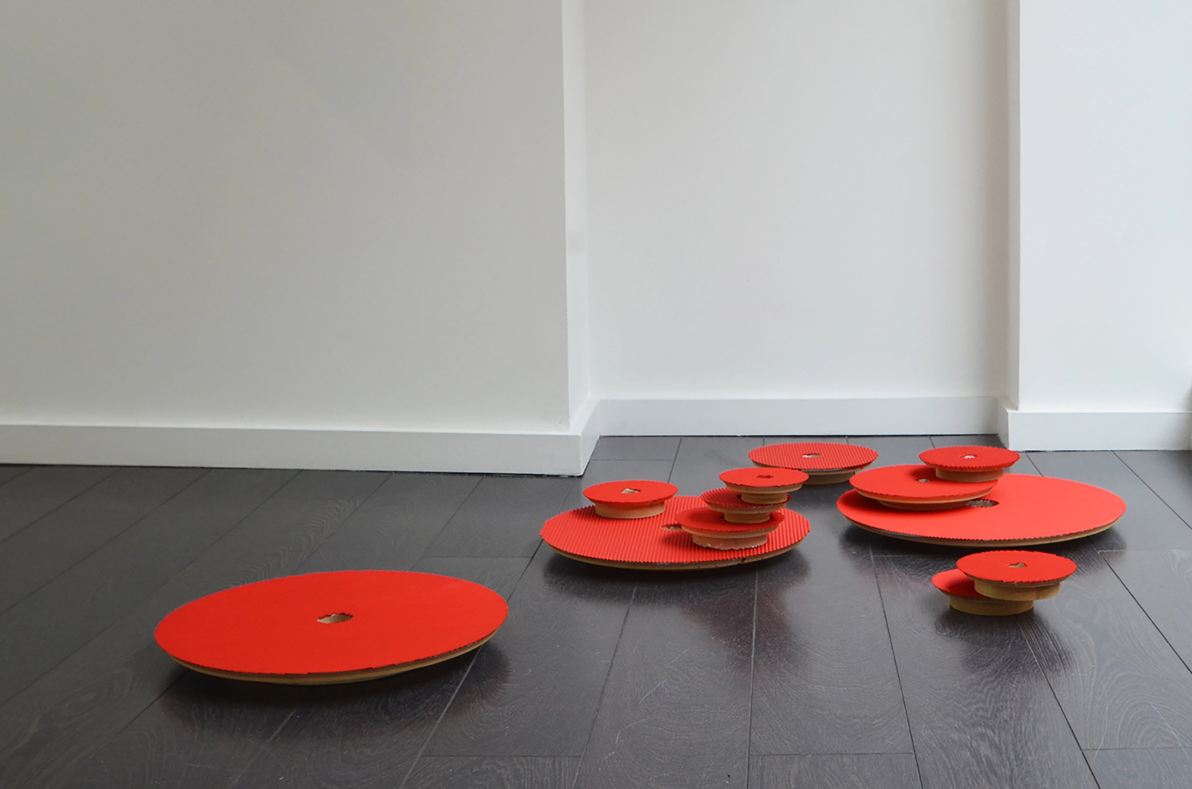 Bright red circular wooden discs of varying sizes arranged across a dark floor, some stacked and tilted, creating a sculptural installation in a gallery space