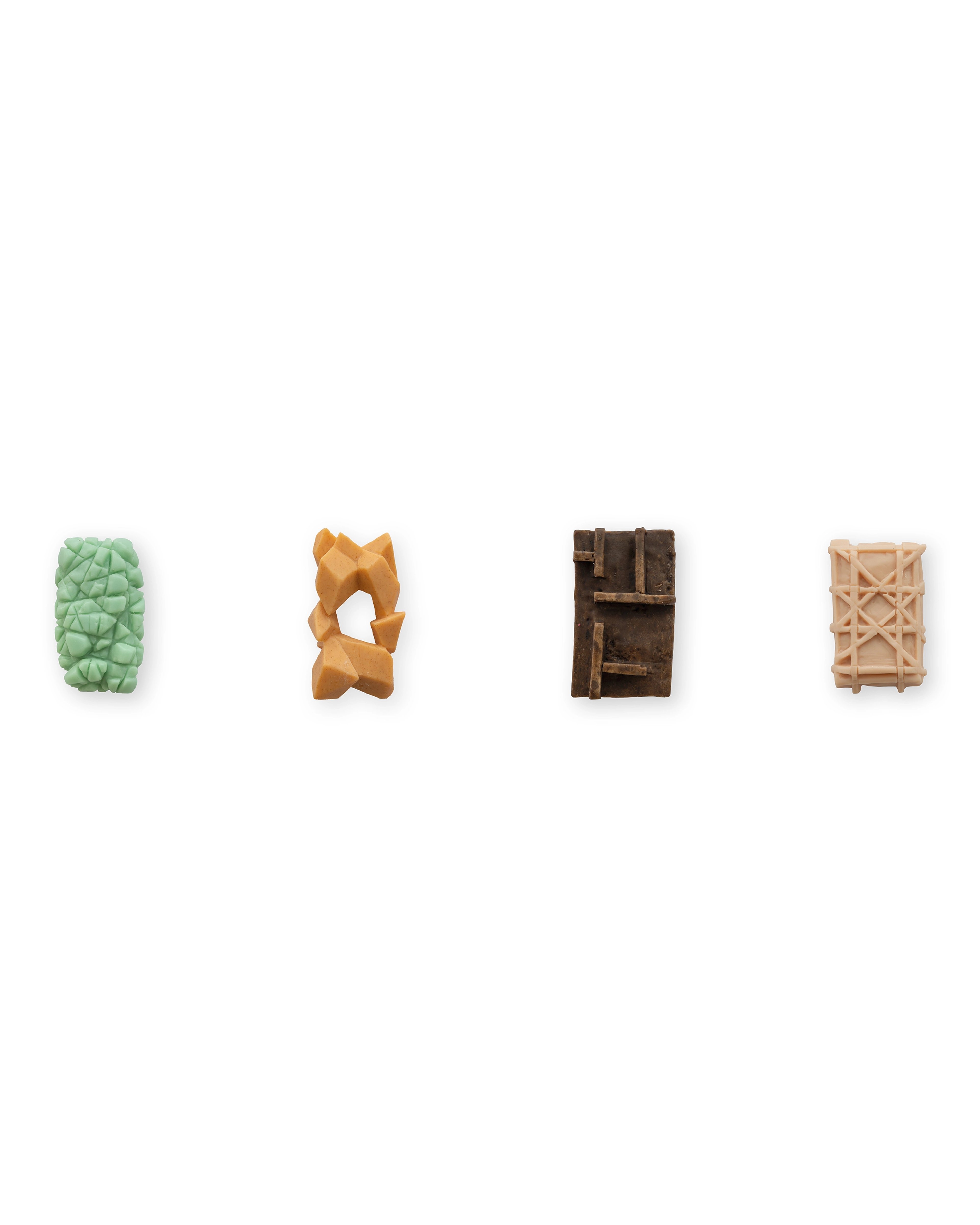 Four carved soap bars in green, orange, dark brown, and pale wood tones arranged in a row, each with distinct textured and geometric forms against a white ba…