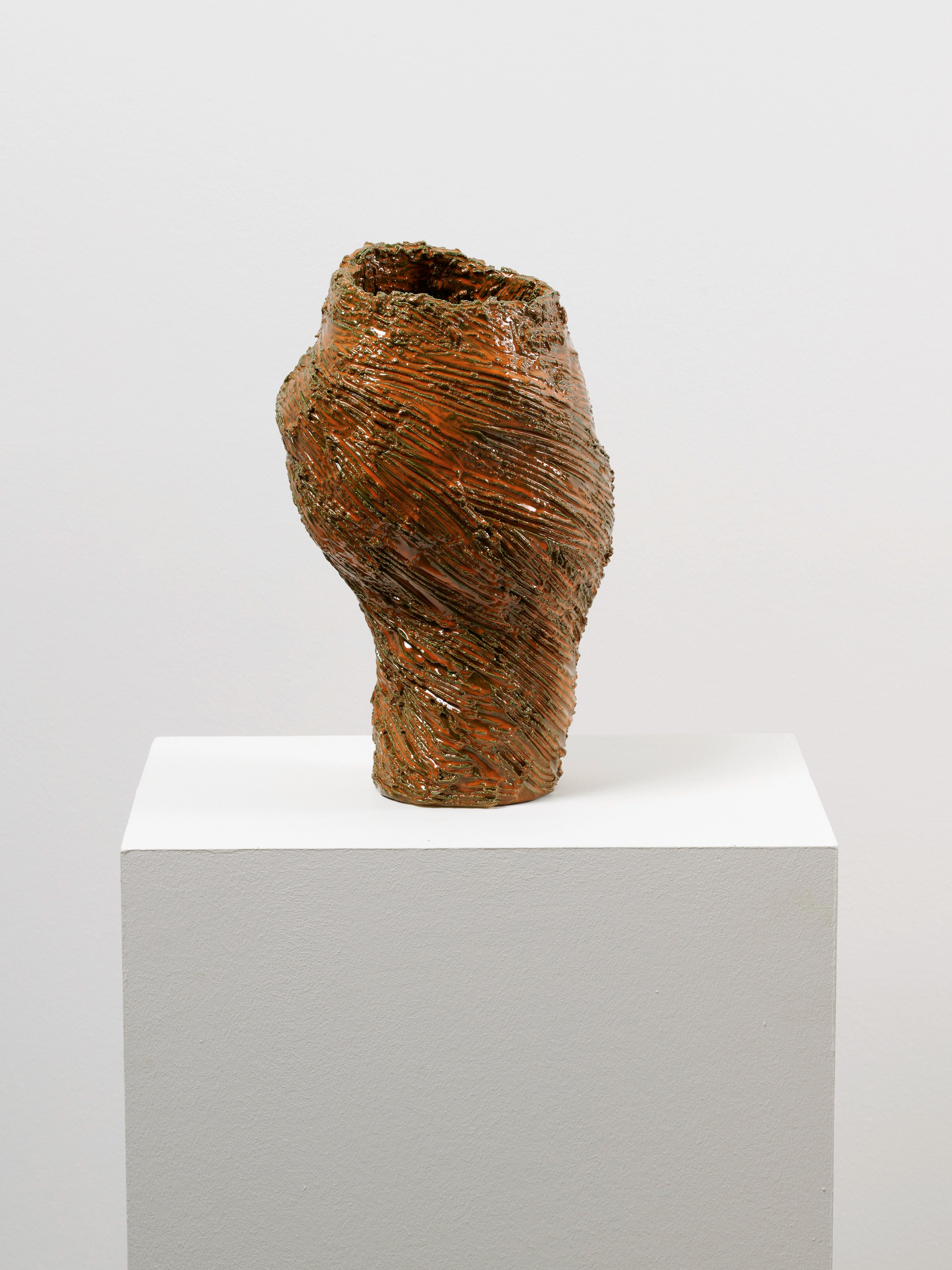 Twisted ceramic vessel with horizontal striations in ochre and burnt orange, mounted on a white plinth against neutral background.