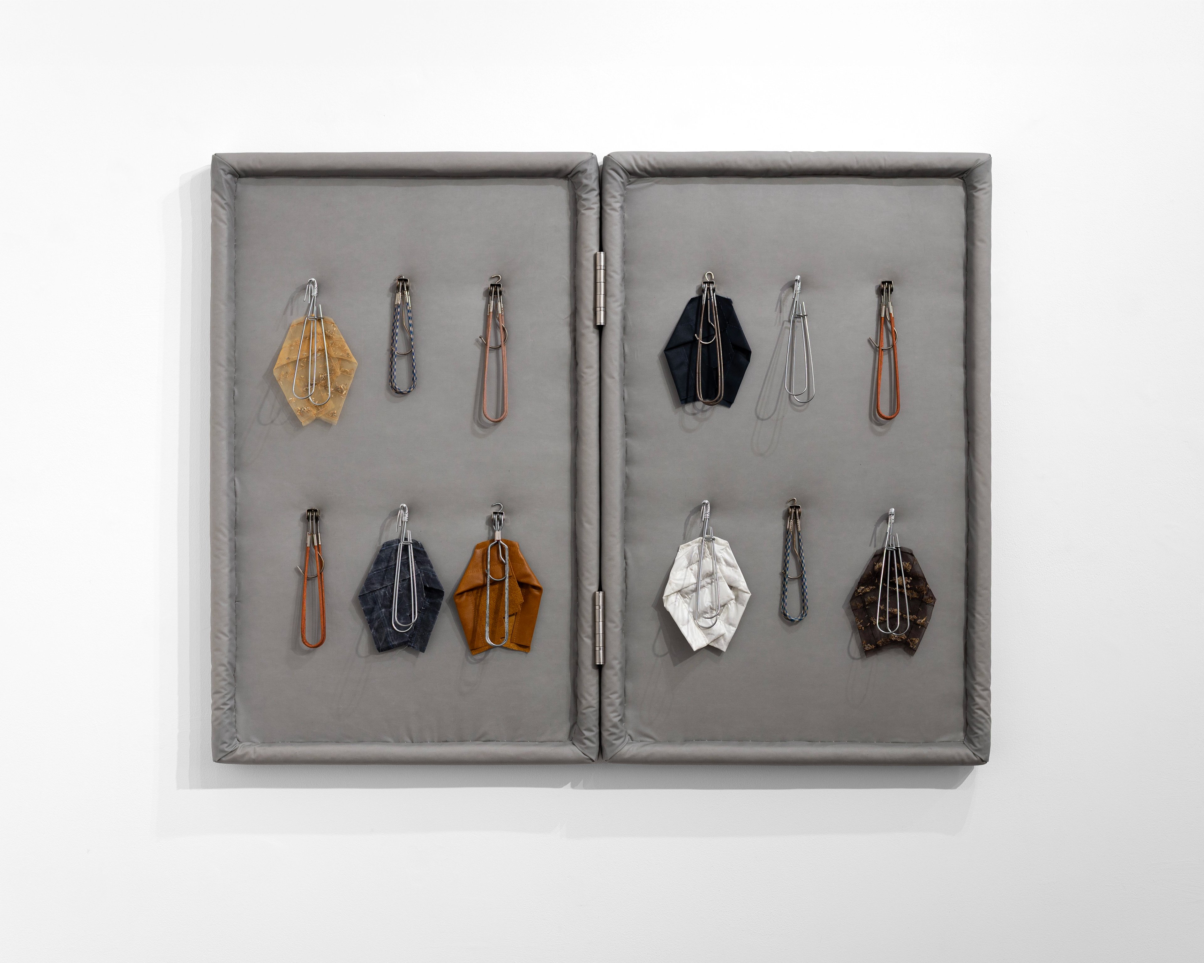 Two gray fabric-lined display cases side by side containing twelve small fabric pouches in earth tones, navy, white, and orange, each suspended from metal ha…