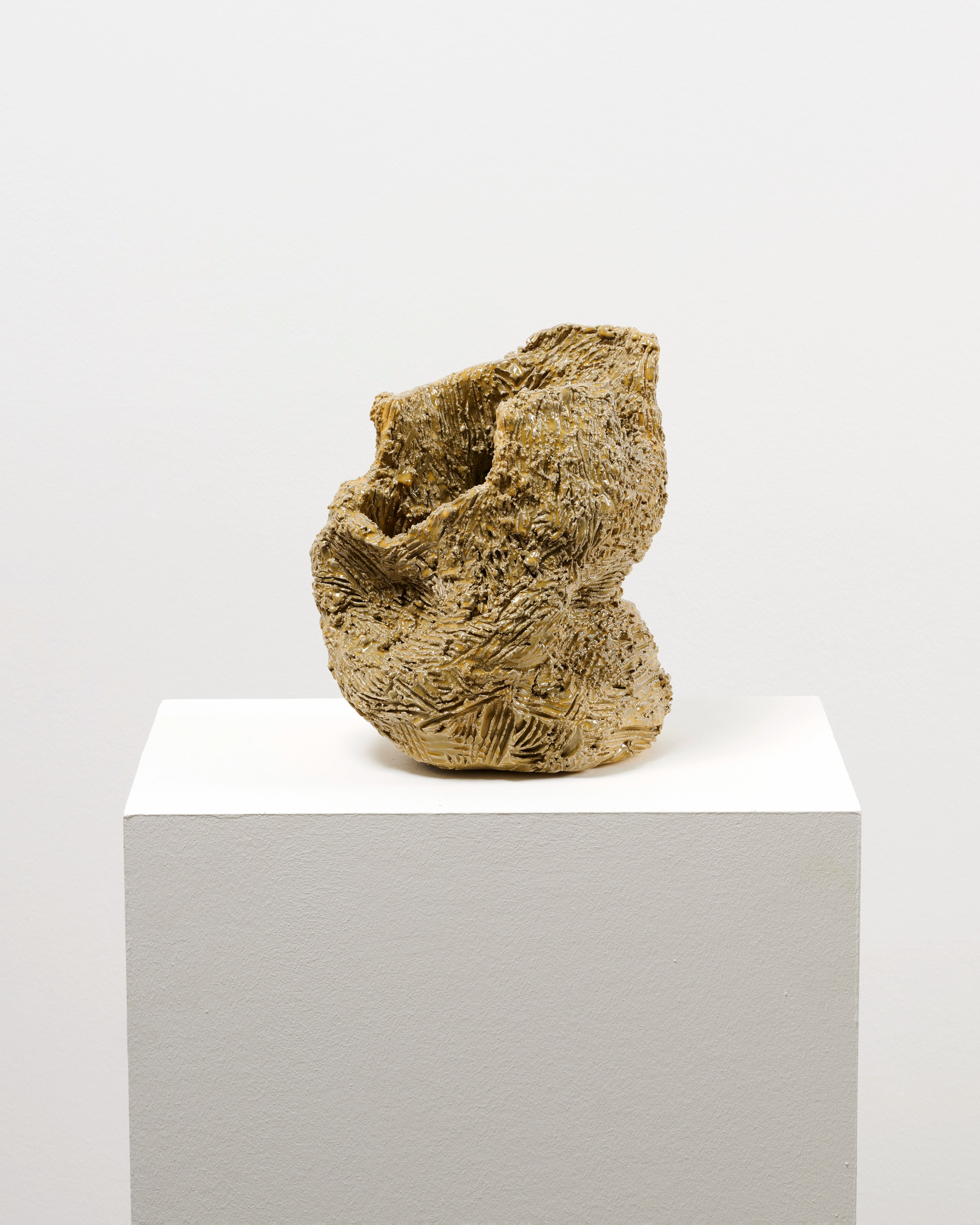 Glazed ceramic sculpture in gold tones with rough, textured surface resembling natural geological forms, mounted on a white-topped grey plinth against a neut…