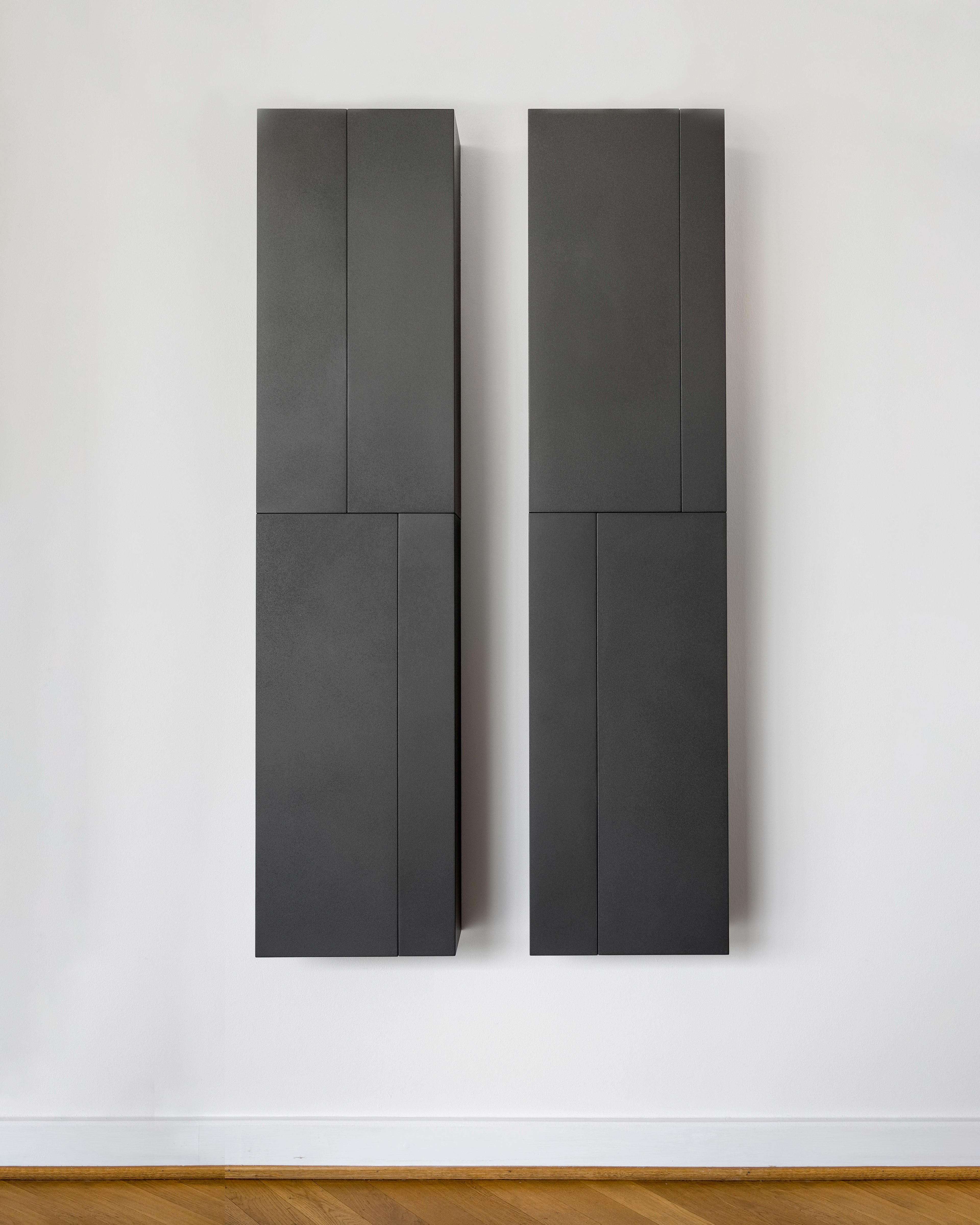 Two tall charcoal-grey wooden panels with vertical divisions mounted side by side on a white wall, their matte surfaces created with pigments, lacquer and wax