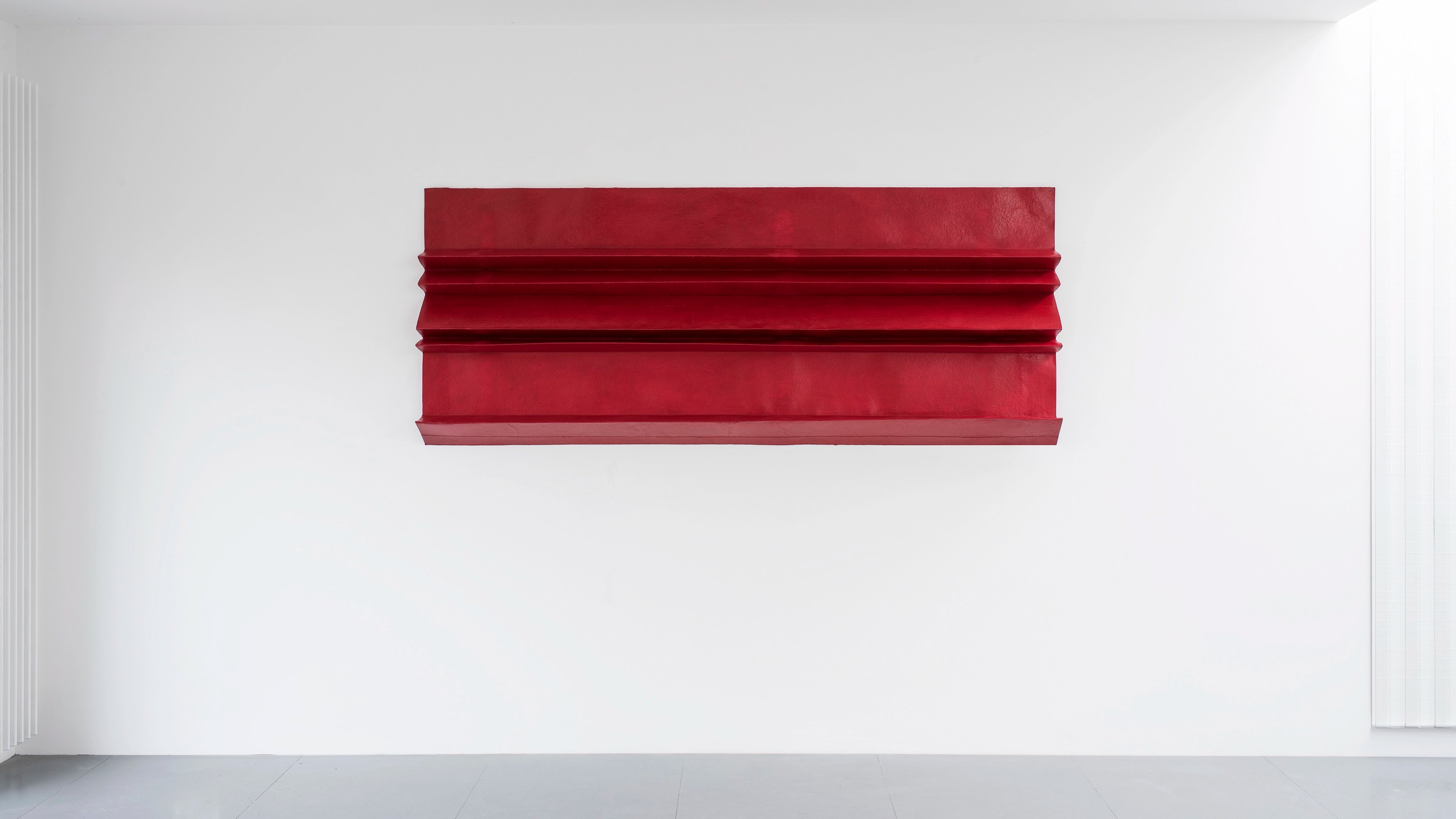 Deep red horizontal paper form with layered, folded sections creating dimensional ridges against white wall