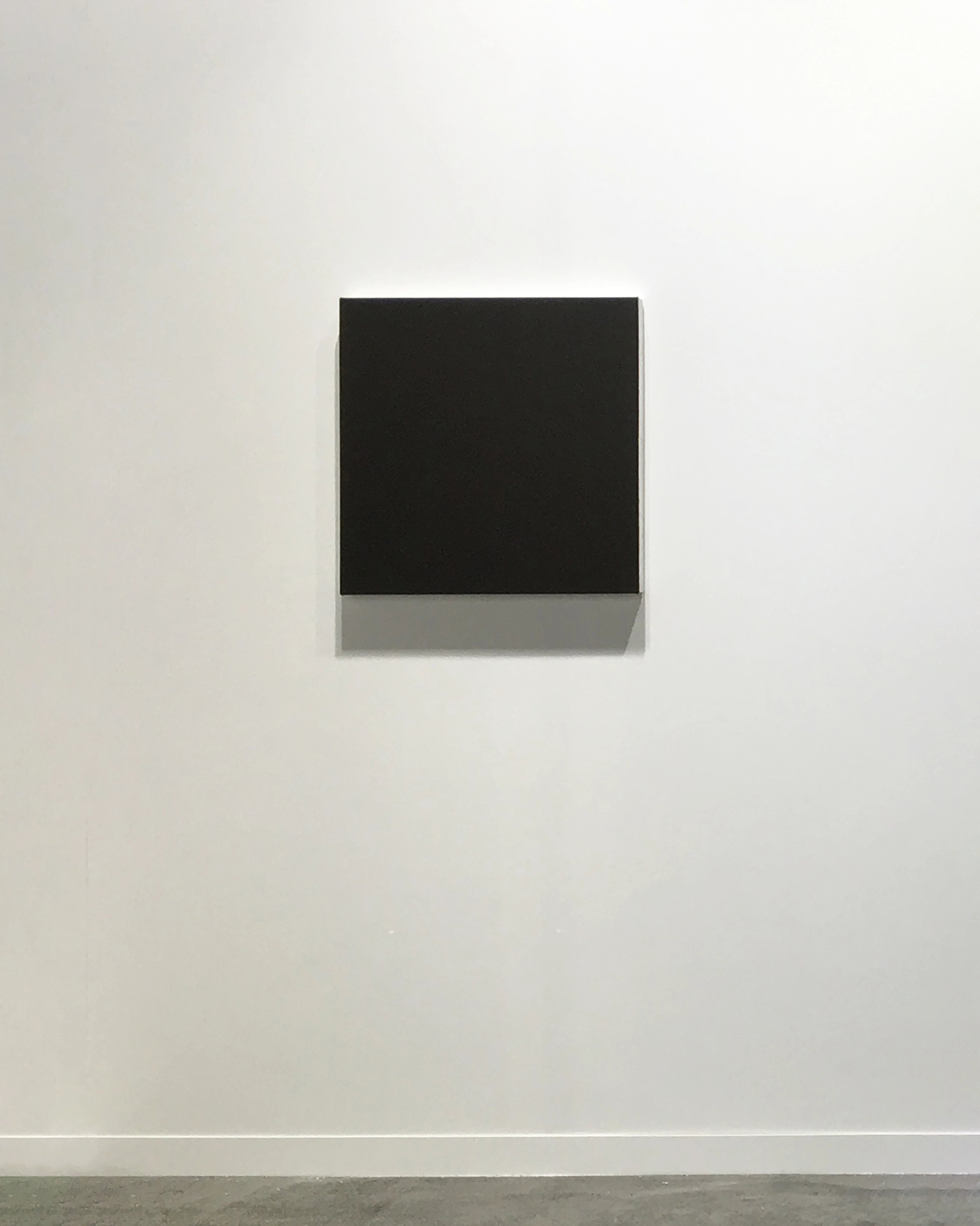 Black square canvas with subtle grey shadow beneath, mounted on white gallery wall