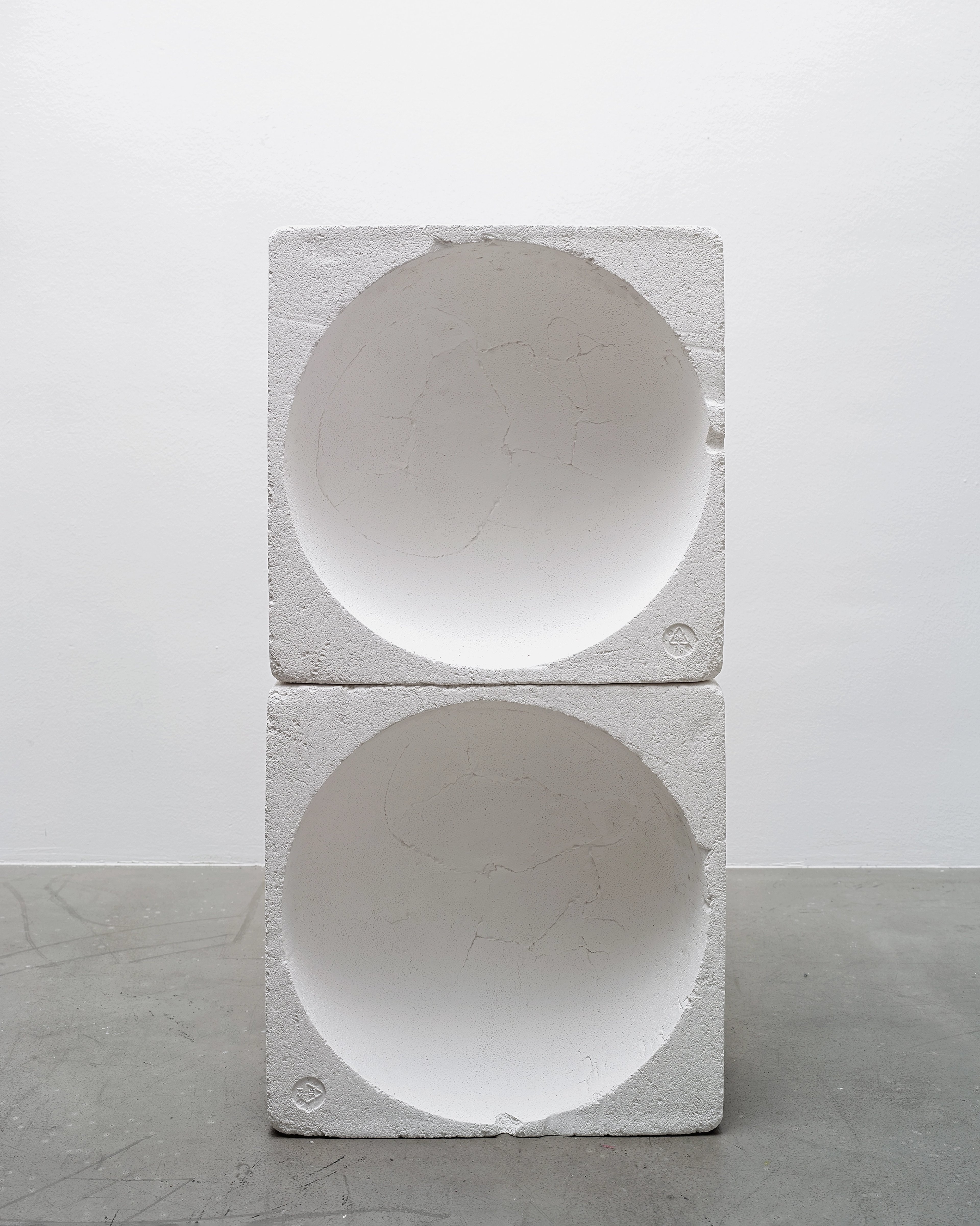 Two stacked white plaster or concrete blocks, each carved with a deep hemispherical hollow, creating mirror-like concave forms against a gallery wall