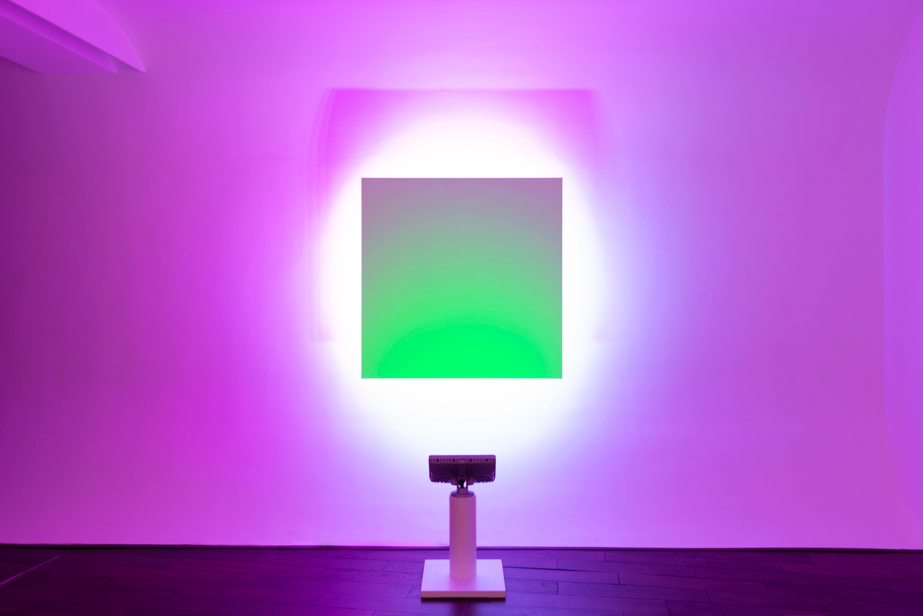 Glowing square LED panel transitioning from grey to vibrant green, mounted on a purple wooden plinth, set against magenta gallery walls suffused with soft li…