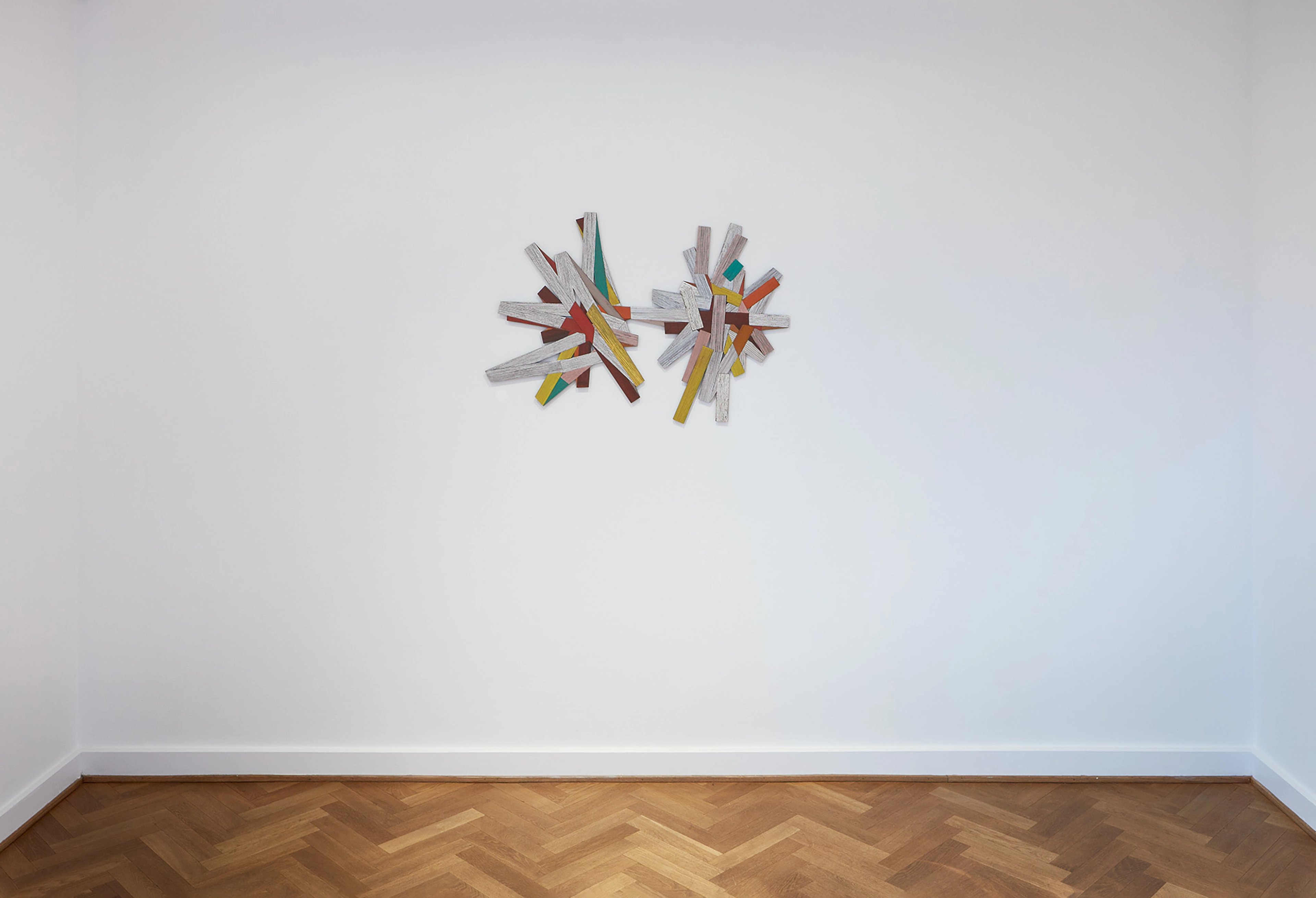 Two radiating clusters of painted wooden sticks in yellows, reds, greens, and whites mounted on a white gallery wall above herringbone parquet flooring
