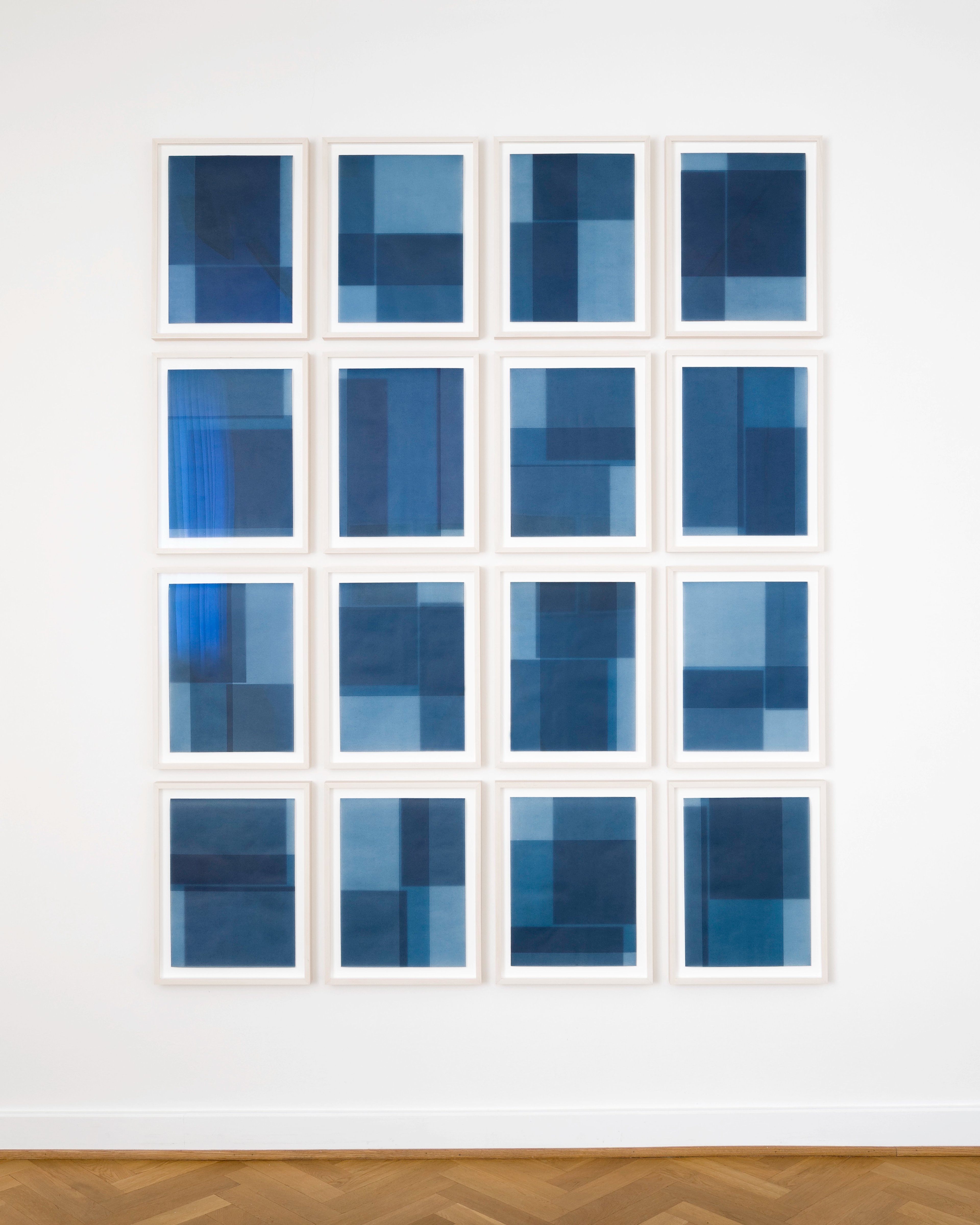 Sixteen framed works on paper in a four-by-four grid, each featuring geometric compositions of overlapping rectangles in shades of blue, from navy to light c…