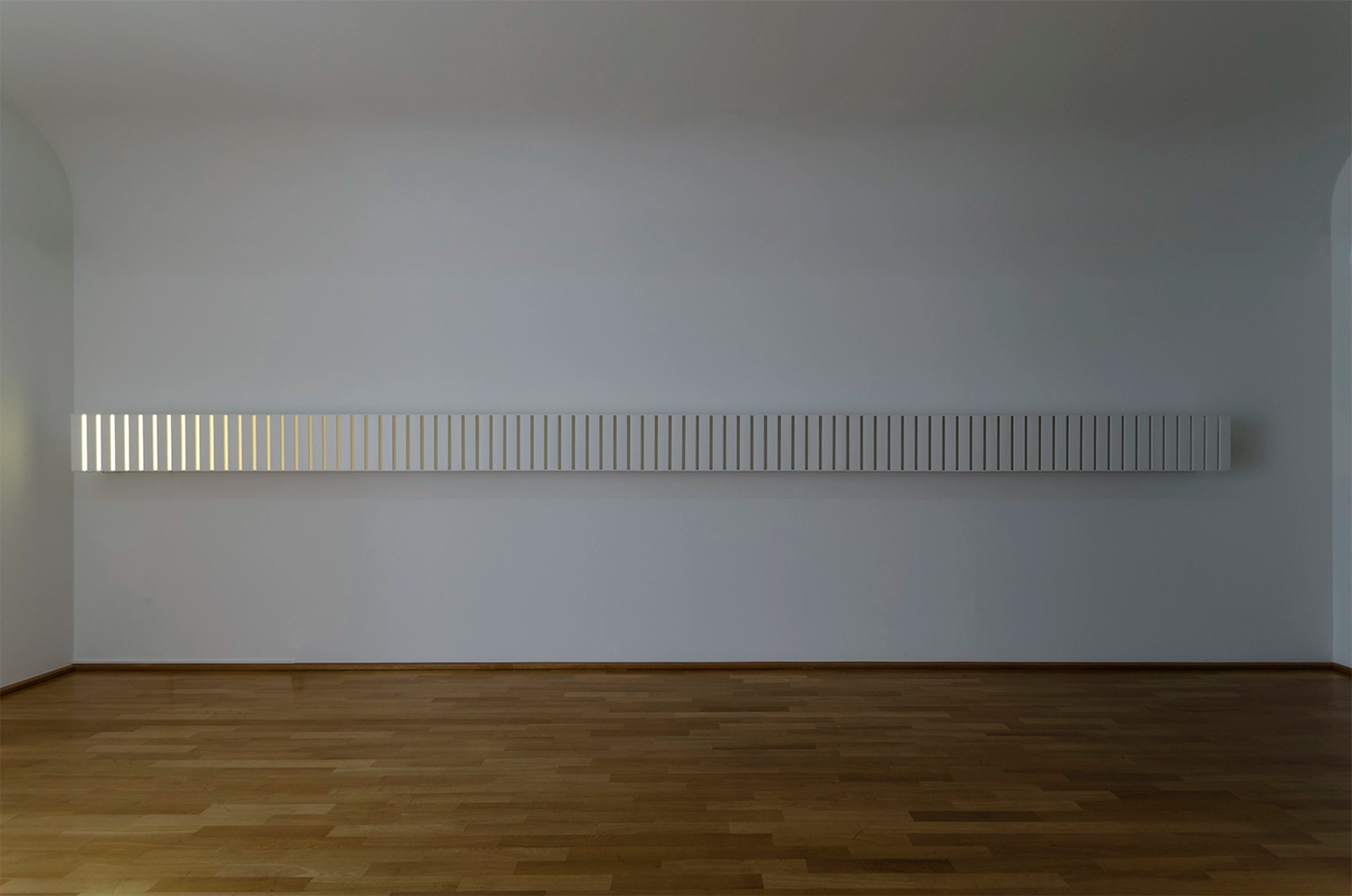 Horizontal installation of evenly spaced white vertical slats with integrated LED lighting mounted on gallery wall above wooden floor