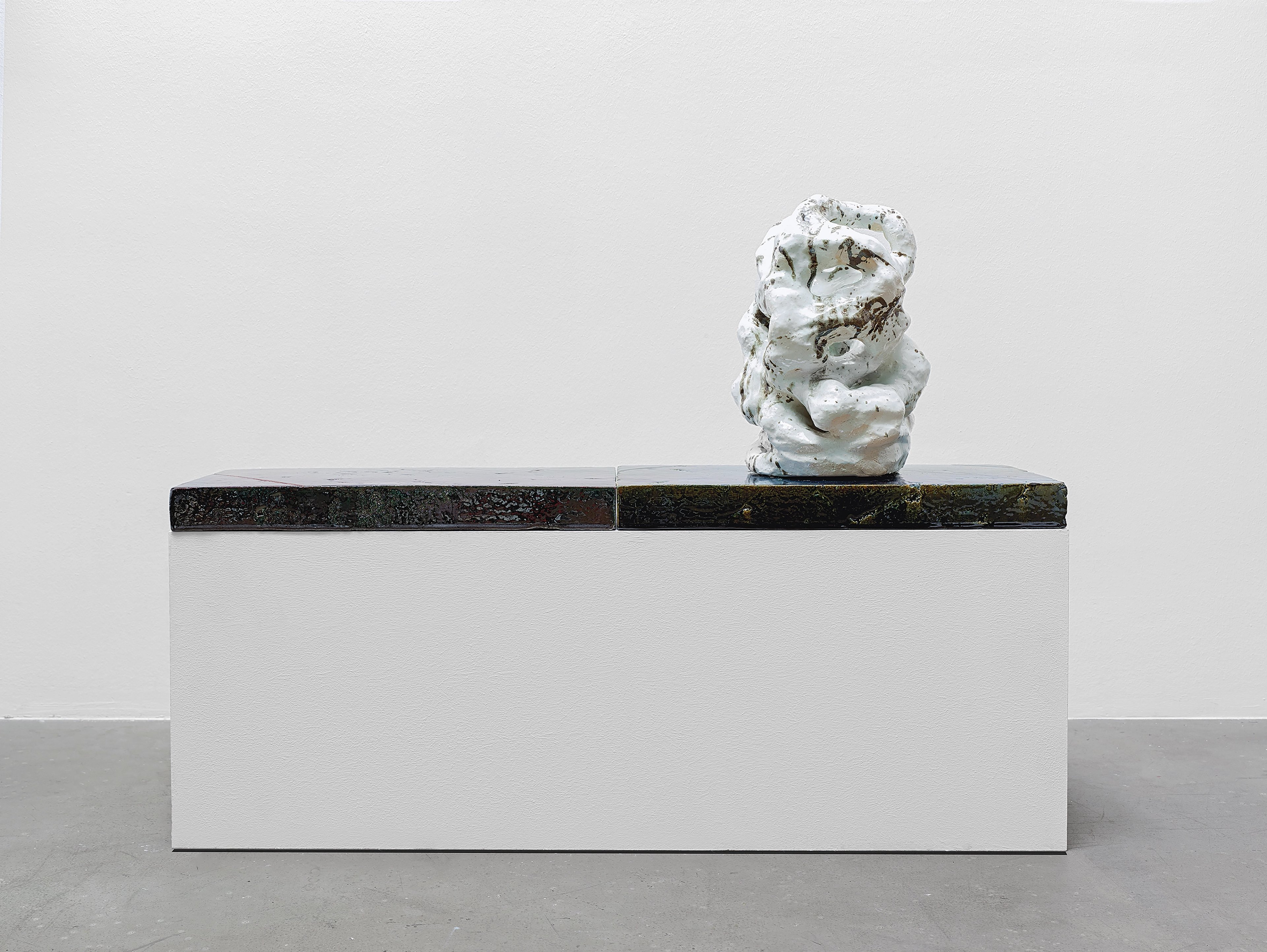 White ceramic sculptural form with organic, crumpled surfaces perched on dark wooden beam atop minimalist white plinth