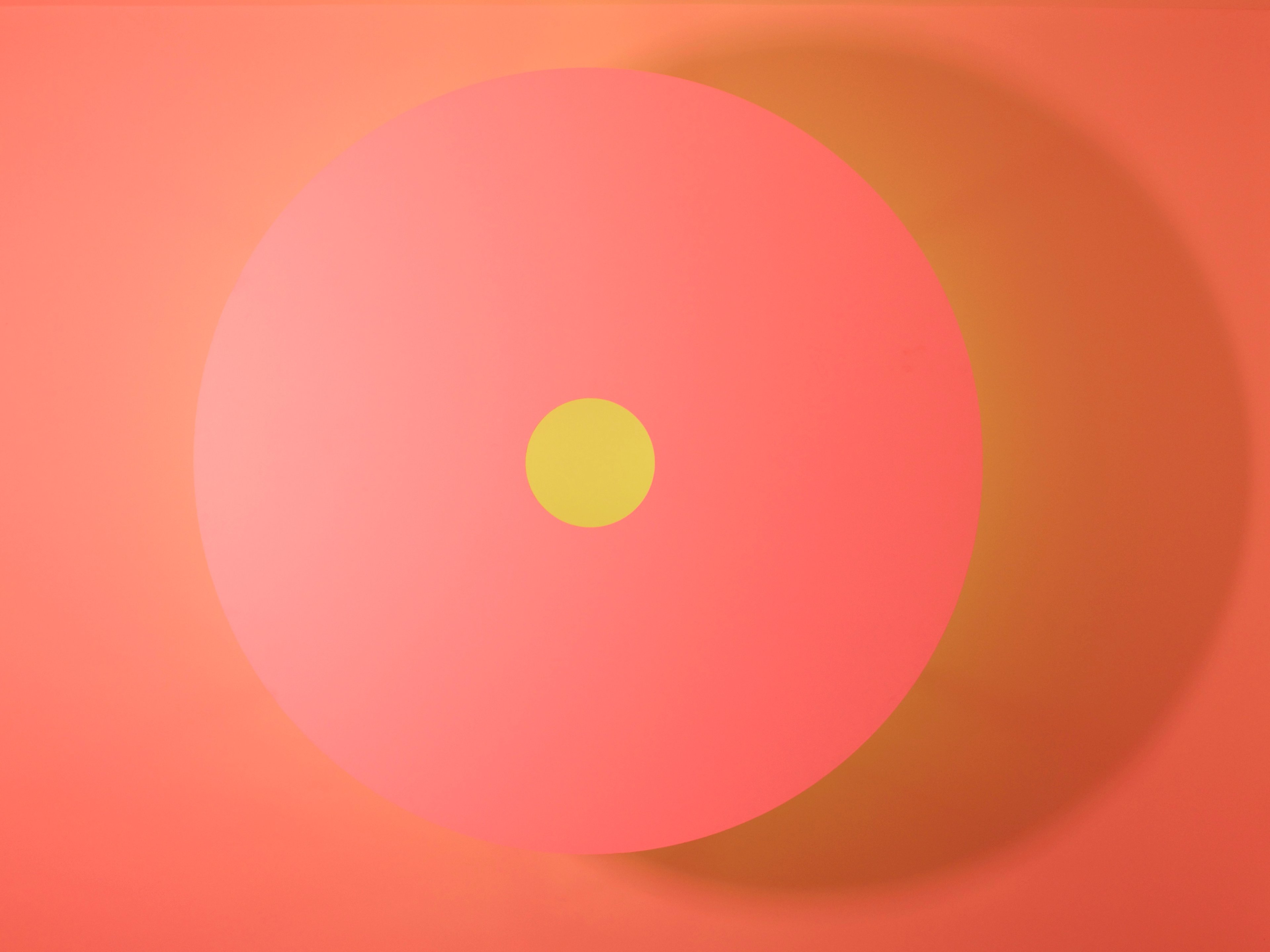 Large coral-pink circular panel with a small yellow circle at centre, casting a warm shadow on the peachy background, minimalist geometric composition