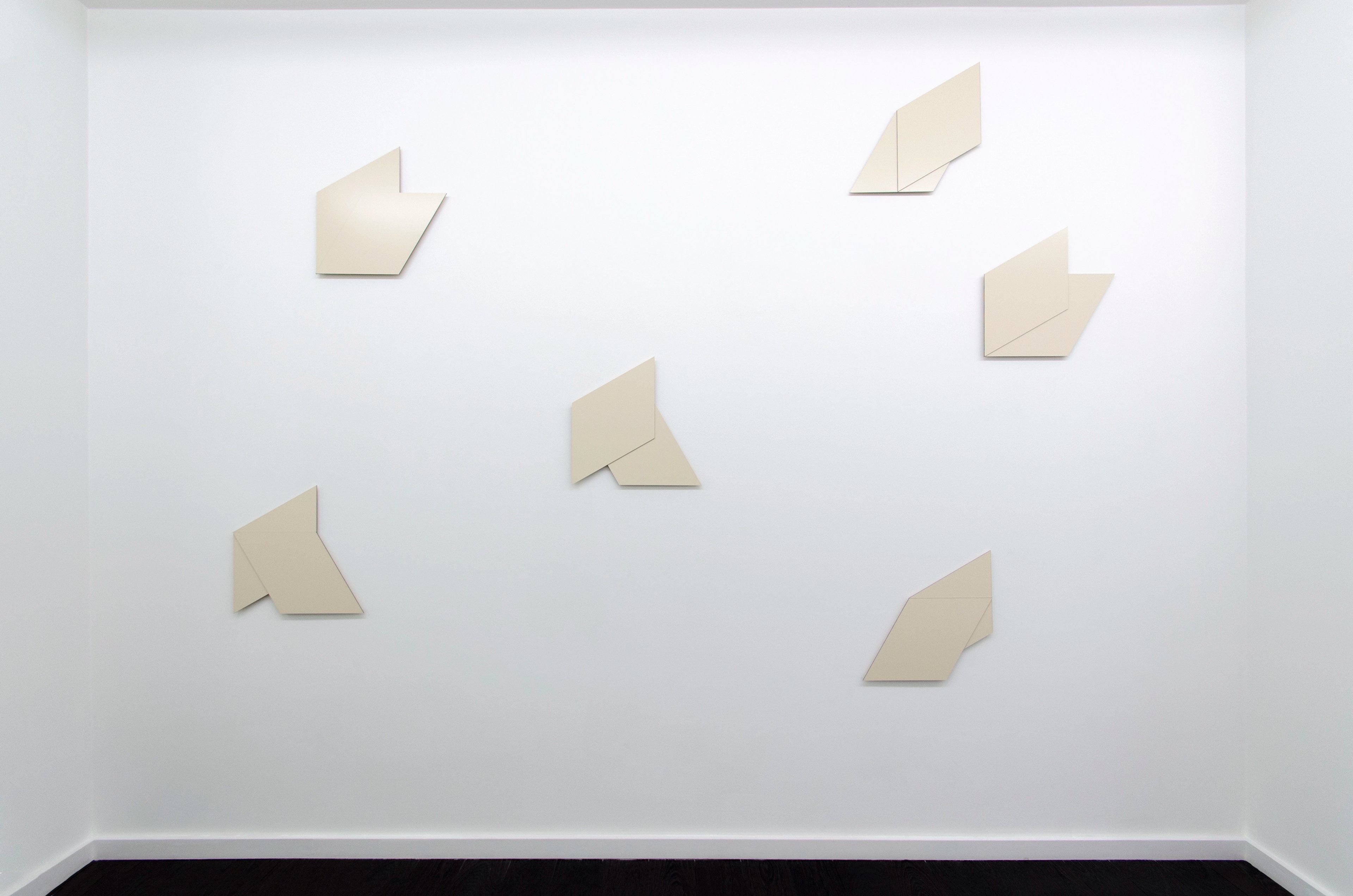 Six cream-coloured geometric wall-mounted panels with angular, folded forms scattered across a white gallery wall