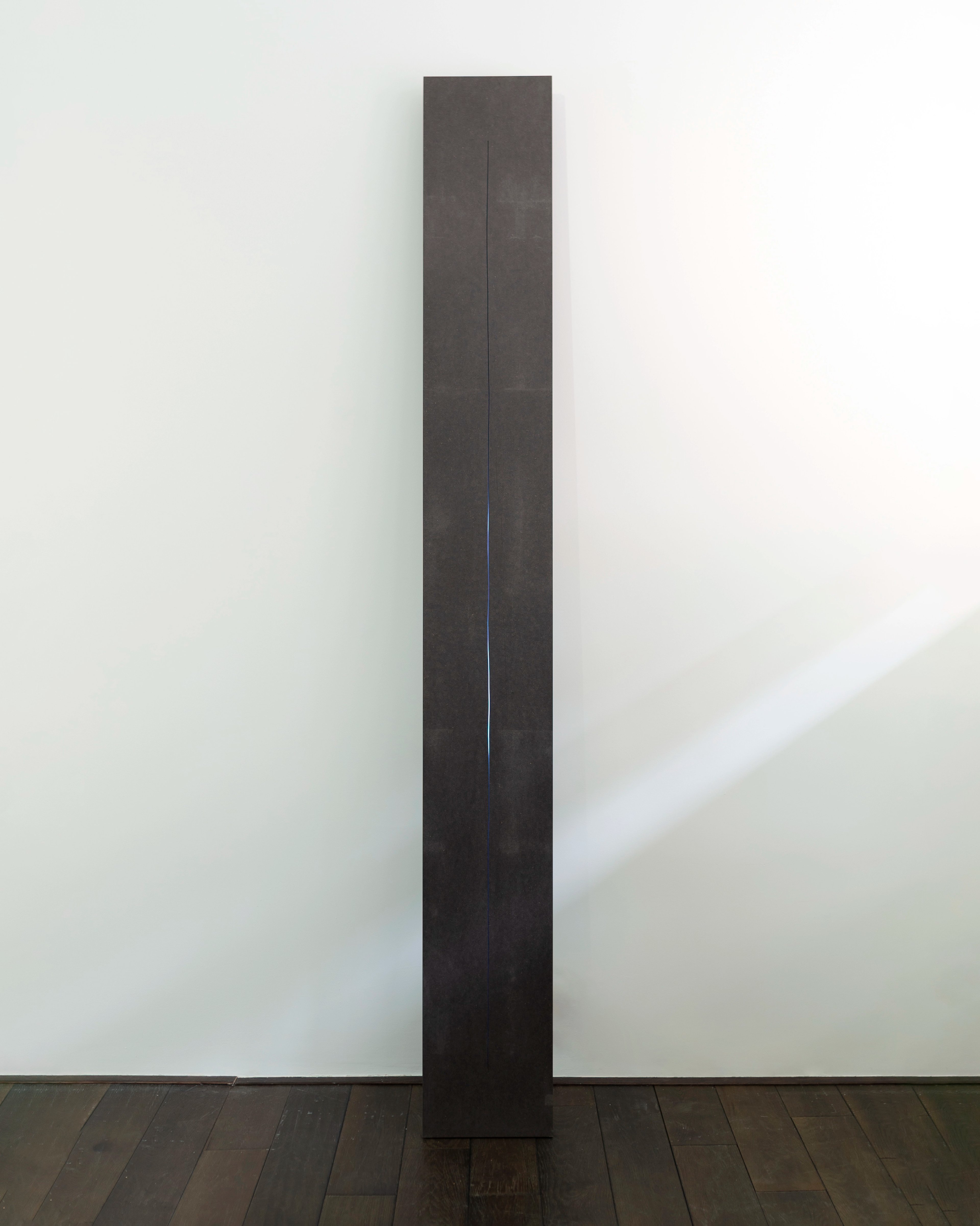 Tall vertical black rectangular column with a thin blue LED light running down its center, mounted on a wooden floor against a white gallery wall