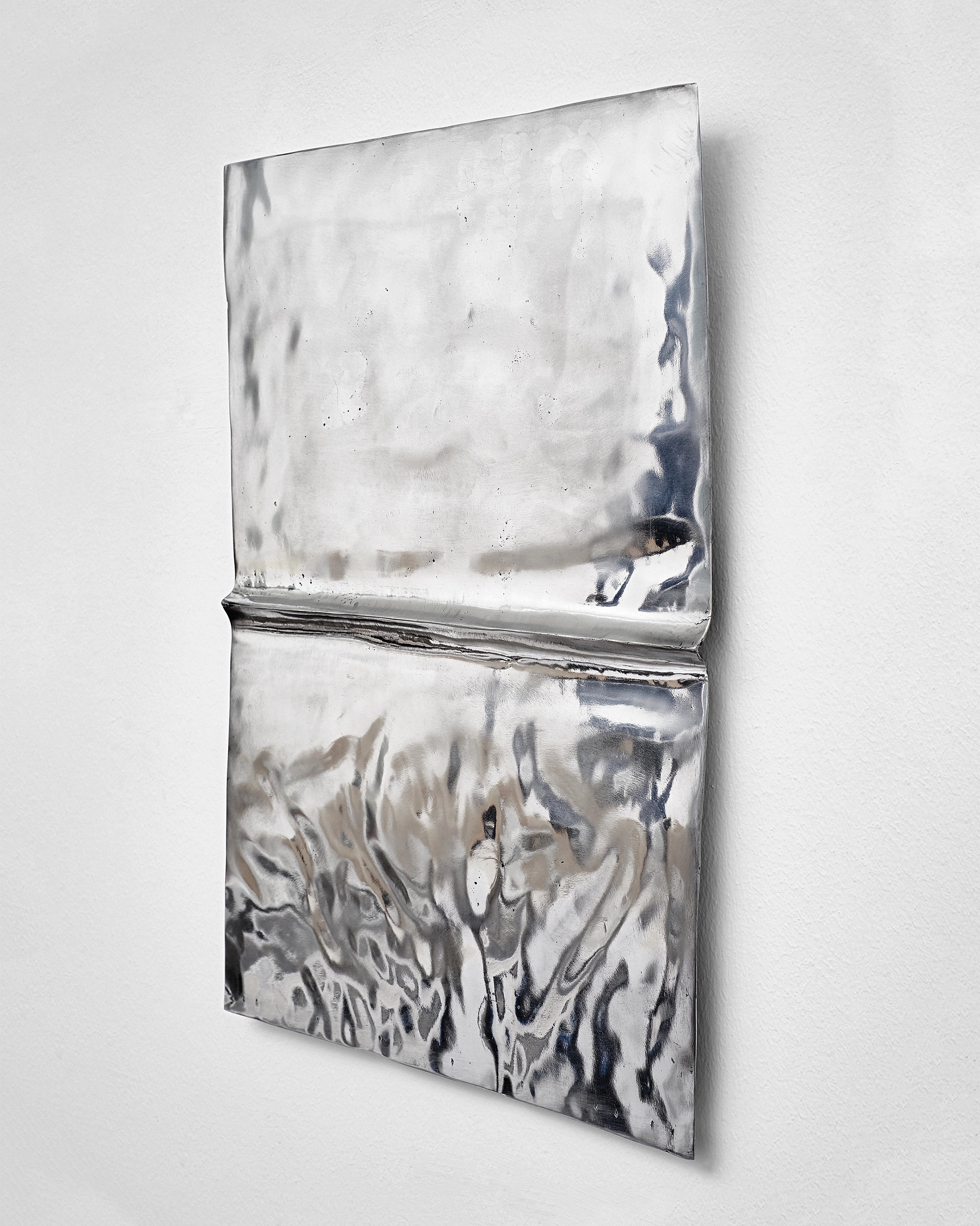 Polished aluminium cast of a folded newspaper mounted vertically on white wall, upper section smooth and reflective, lower section with deeply textured surfa…