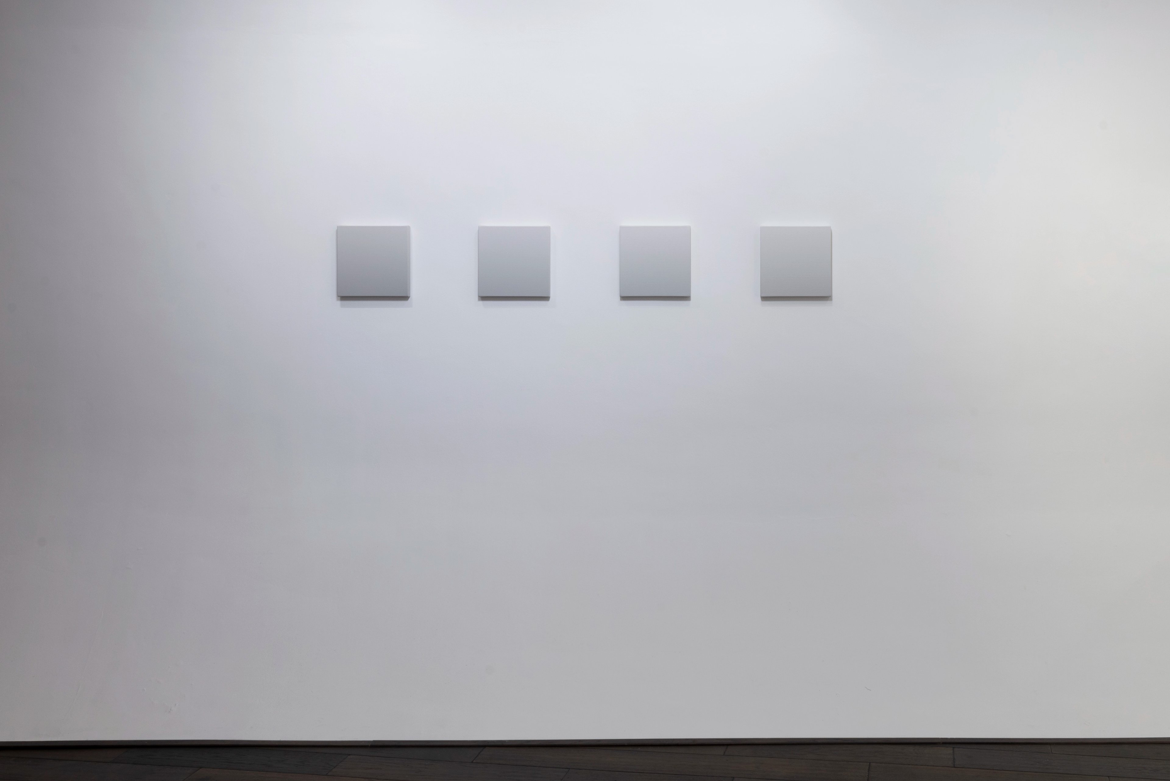 Four gray square canvases mounted horizontally in evenly spaced progression on white gallery wall