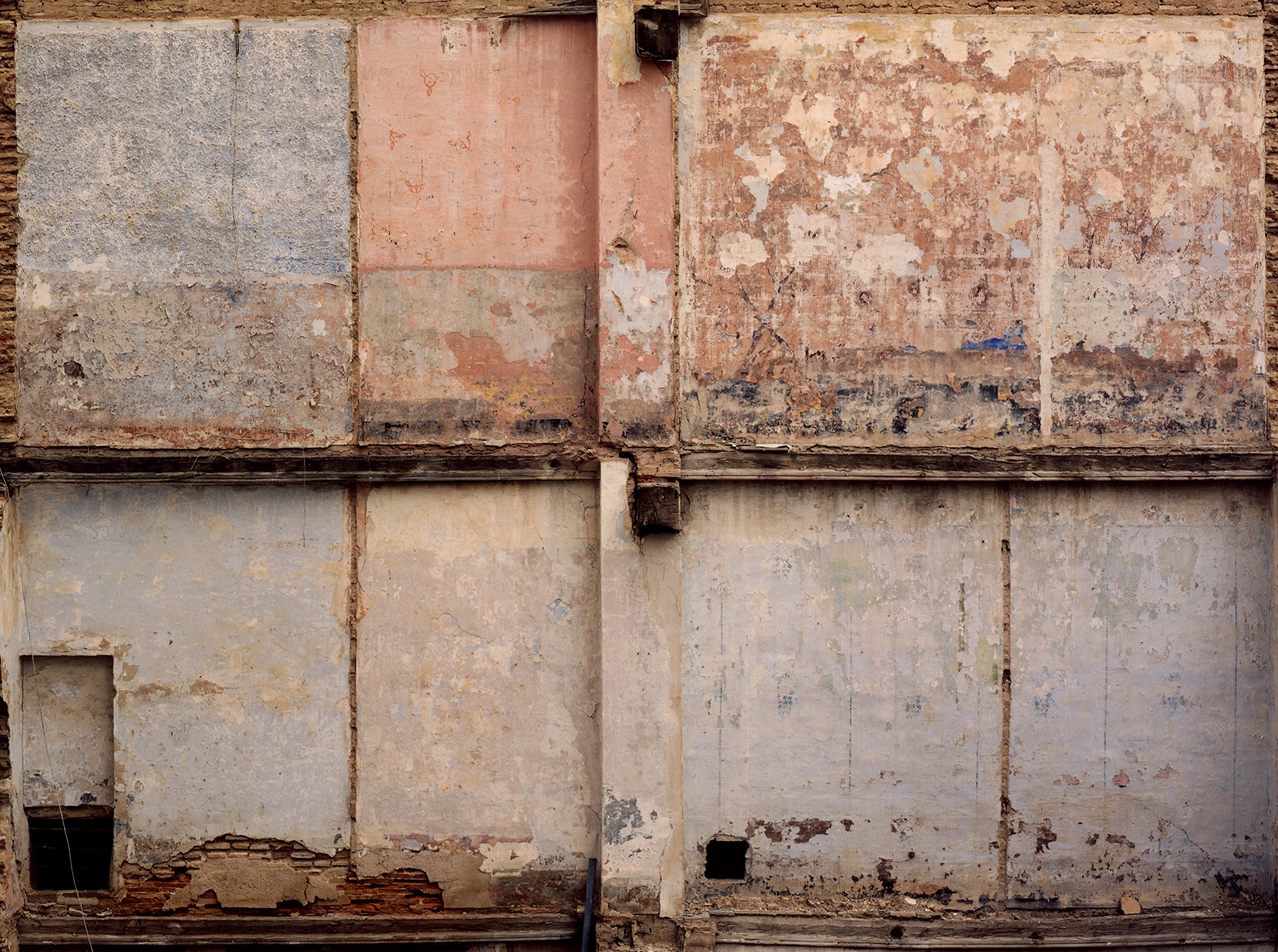 Weathered concrete wall divided into rectangular panels in rust, cream, and grey tones, with peeling paint, stains, and small rectangular openings suggesting…