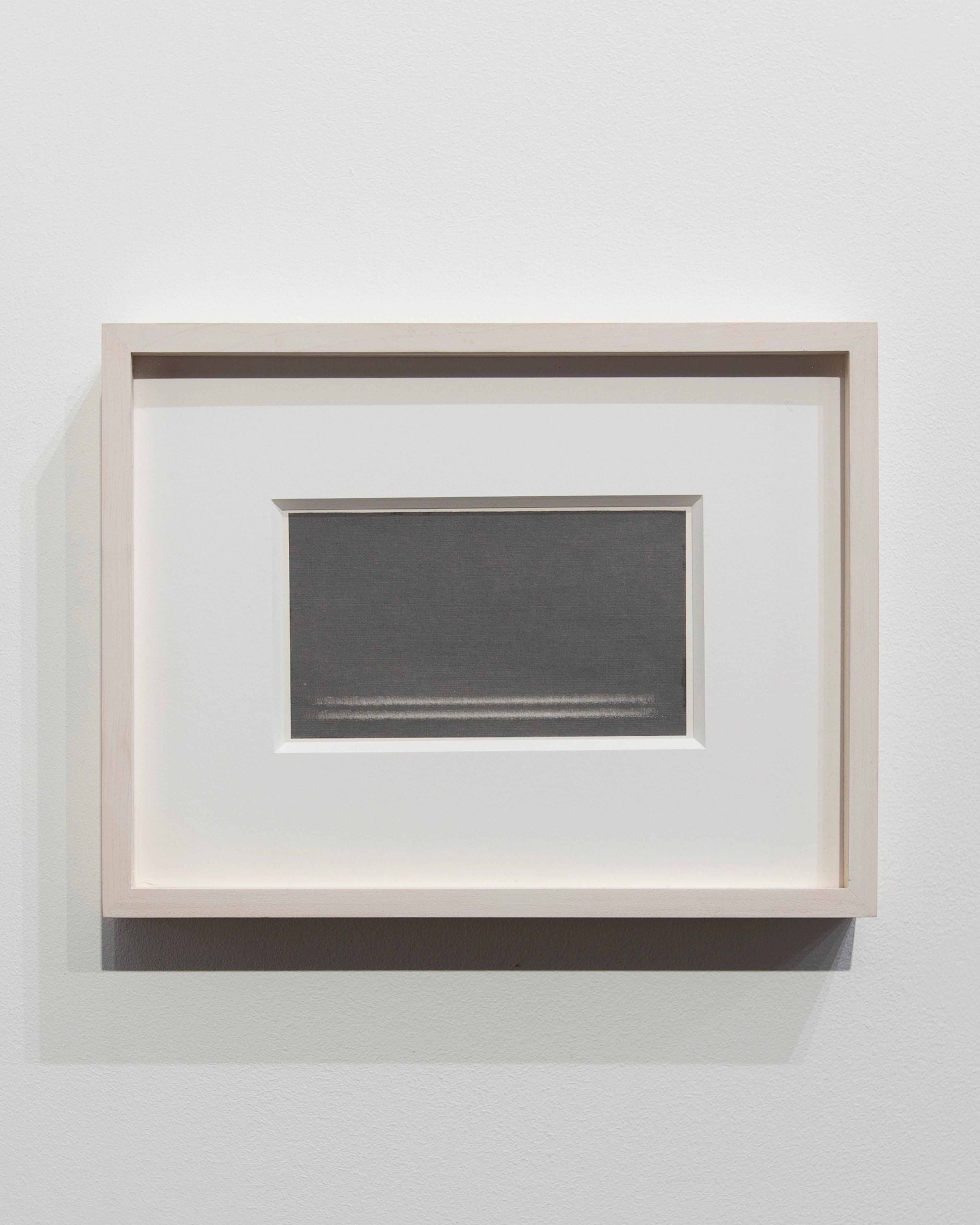 Graphite transfer on rag paper mounted within a light wooden frame with dark horizontal bands, depicting a minimal rectangular dark form centered on white gr…