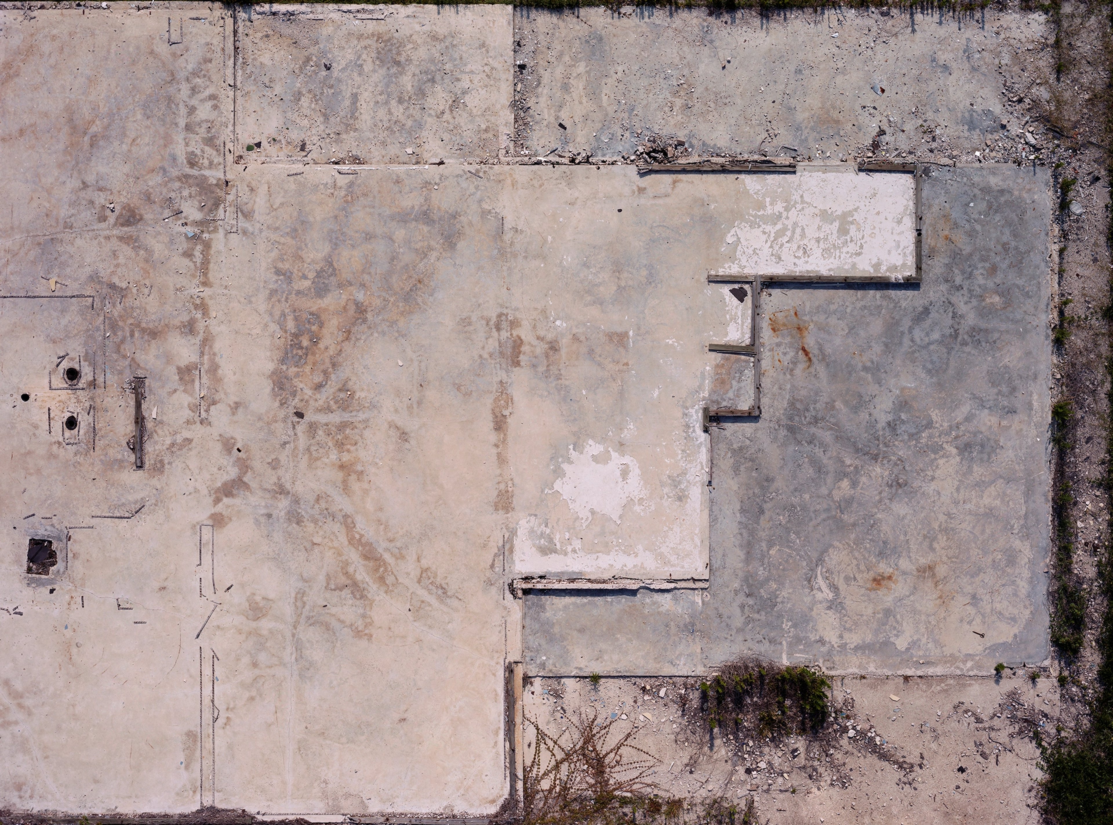 Aerial view of weathered concrete pavement with faded rectangular sections, visible seams, and sparse vegetation growth along edges