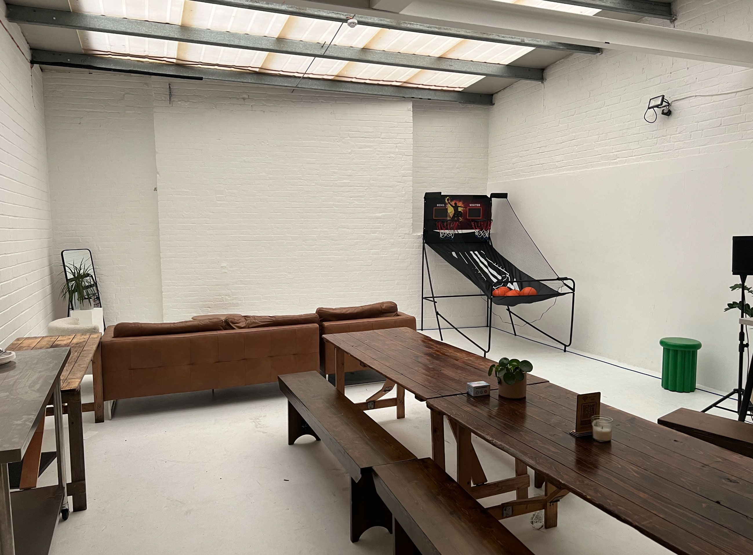 Interior view of Brixton Warehouse showing a bright white studio with long wooden dining tables, tan leather sofas, and a basketball hoop game setup — a versatile space for creative shoots and events.