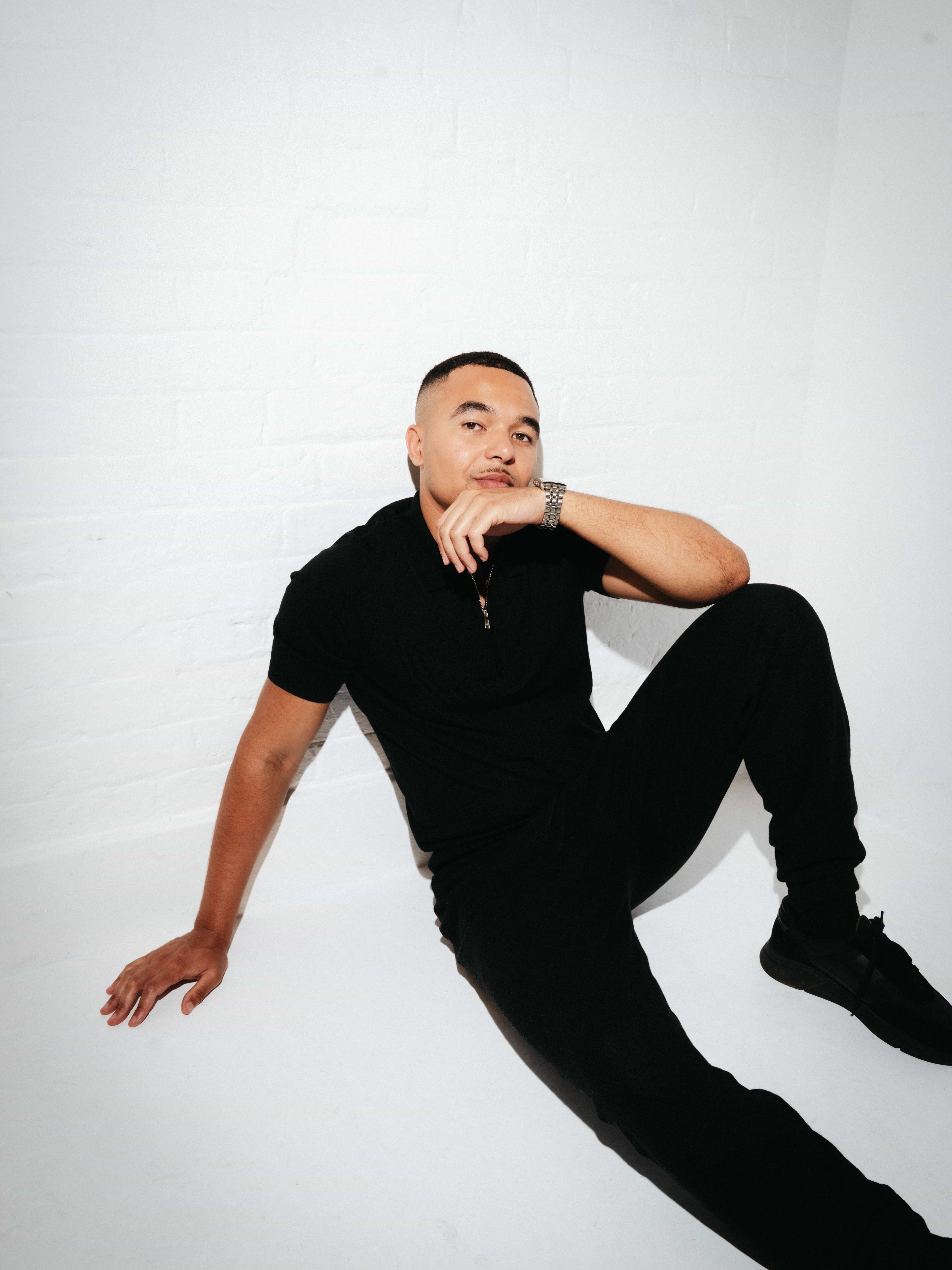 A young man dressed in all black sits casually on the floor against a white brick wall. He rests one arm on his bent knee and the other on the floor, gazing thoughtfully at the camera. He wears a short-sleeved polo shirt, joggers, black trainers, and a silver watch. The setting is minimalist and brightly lit.          Ask ChatGPT