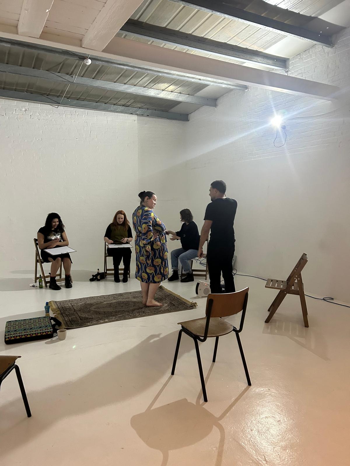 "Life drawing class at Brixton Warehouse in South London. Participants sit with sketchbooks in a bright, industrial-style studio while models pose on a rug in the center of the room. The creative workshop offers a welcoming space for beginners and experienced artists to practice figure drawing and connect through art."