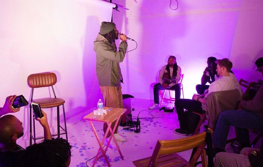 Man holding a microphone surrounded by a crowd at a spoken word, poetry, and music event in a warehouse setting.