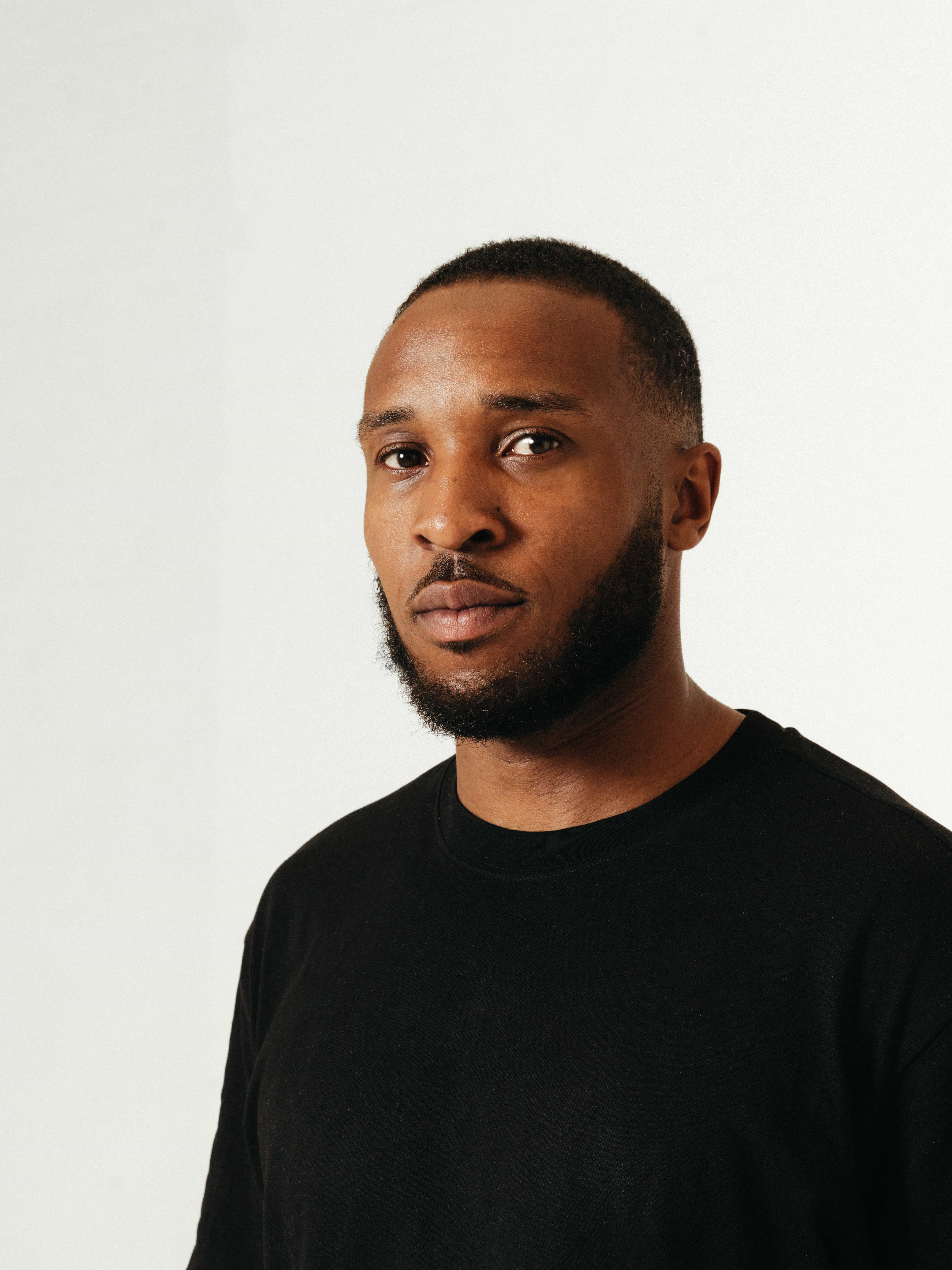  Close-up portrait of a man with a short beard and close-cropped hair, wearing a plain black t-shirt, standing against a neutral white background. Professional headshot of a Love Locations team member in a minimalist studio setting.