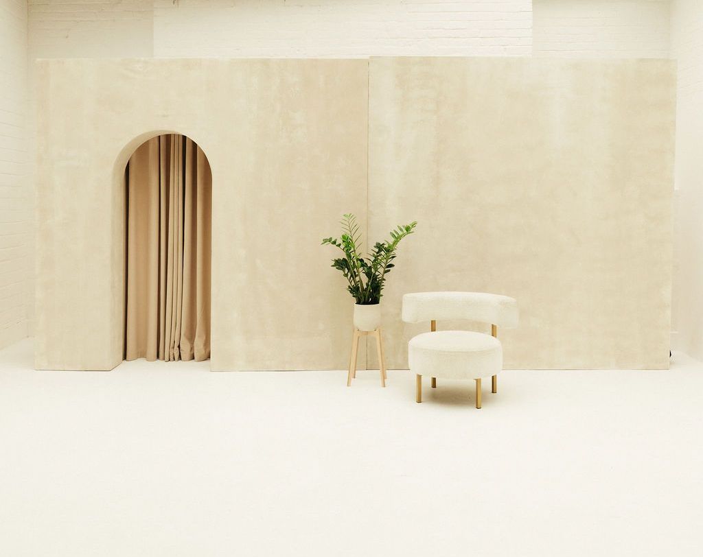 A bright, minimalist area inside Brixton Warehouse featuring a soft cream backdrop with an arched curtain opening, a modern cream chair with gold legs, and a potted plant. The clean, neutral-toned space shows the venue’s versatility for photoshoots, events and styled installations.