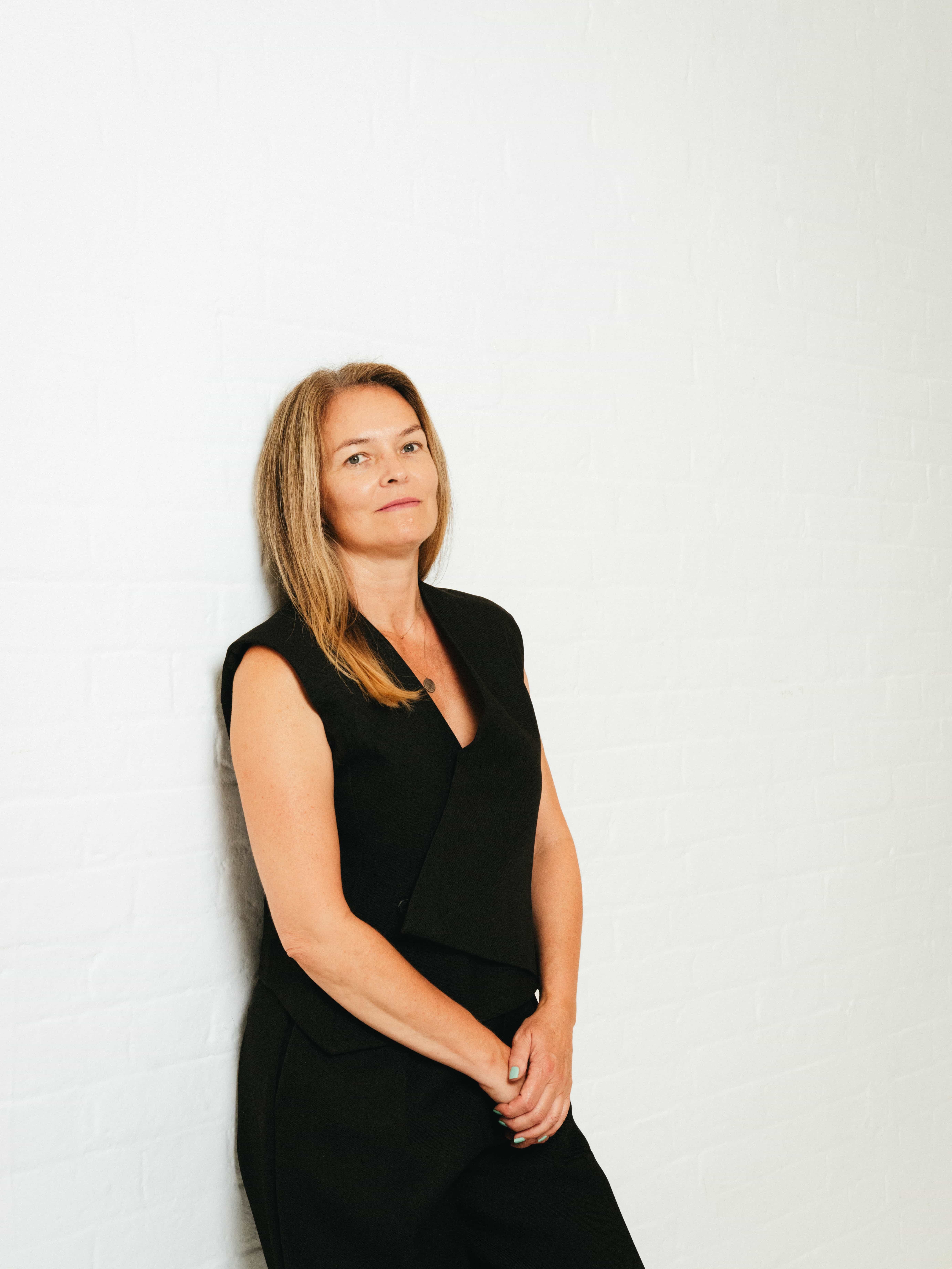 Professional portrait of a woman with straight blonde hair, wearing a black sleeveless outfit, standing against a white brick wall. Styled in a minimalist setting, ideal for creative agency or team bio page. Love Locations team member.