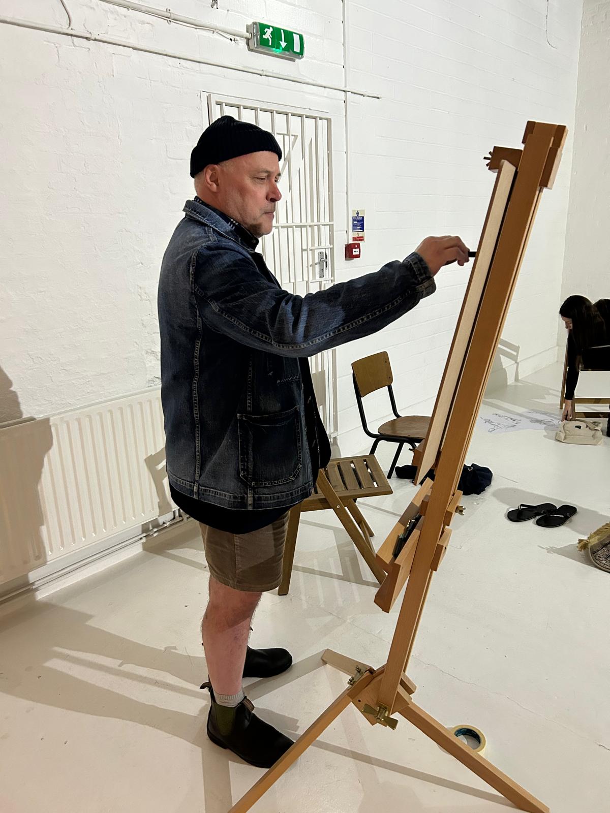 "Artist standing at an easel in a bright white studio during a life drawing class. Wearing a denim jacket, shorts, and a black beanie, he concentrates on sketching while surrounded by chairs and drawing materials in the creative Brixton Warehouse space."