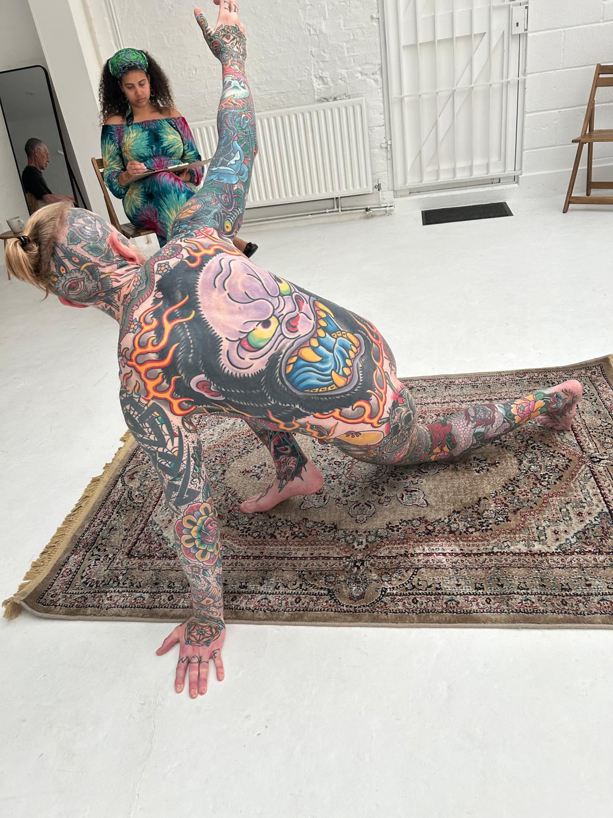 "Life drawing session at Brixton Warehouse featuring a heavily tattooed model posing on a chair and rug in a minimalist art studio. Participants observe and sketch the figure from different angles, creating a dynamic and creative workshop environment in South London."